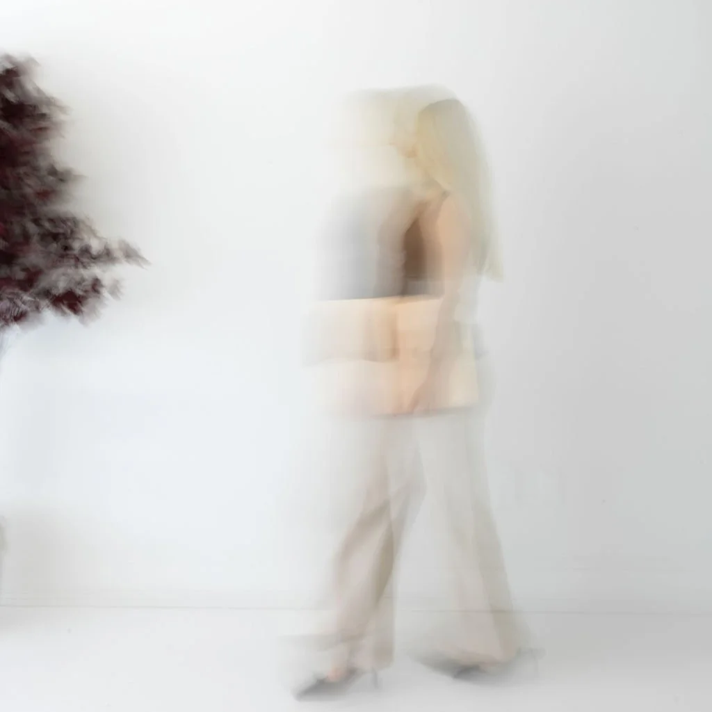 Abstract motion-blurred image of a person walking in front of a plain white wall with a piece of dark-colored foliage on the left side.