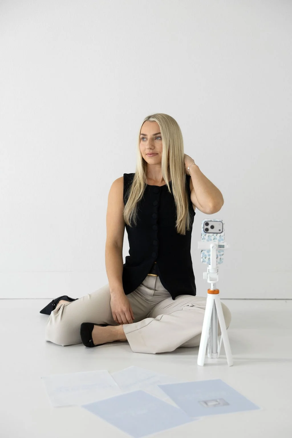Woman sitting on the floor in front of a camera on a tripod, adjusting her hair, with papers on the floor.