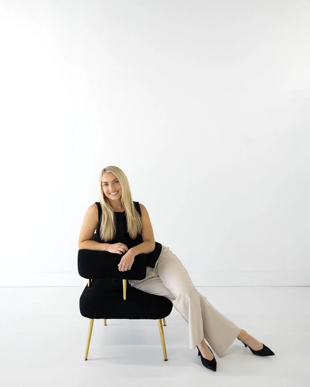 A woman with long blonde hair sitting on a black modern chair with gold legs, smiling, wearing a black sleeveless top, beige wide-leg pants, and black high heels, against a plain white background.