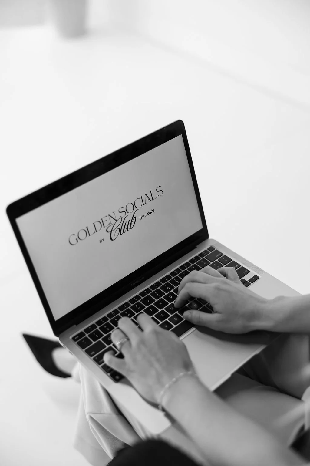 Black and white photo of a person using a laptop with the screen displaying 'GOLDEN SOCIALS CLUB BY BROOKE.' The person is sitting at a white surface, wearing a bracelet.