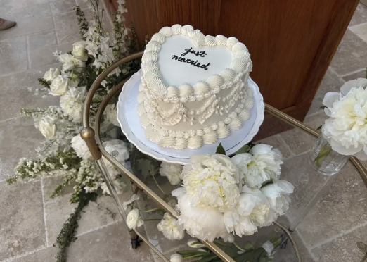A white wedding cake with the words 'just married' on top, placed on a glass cart surrounded by white flowers.