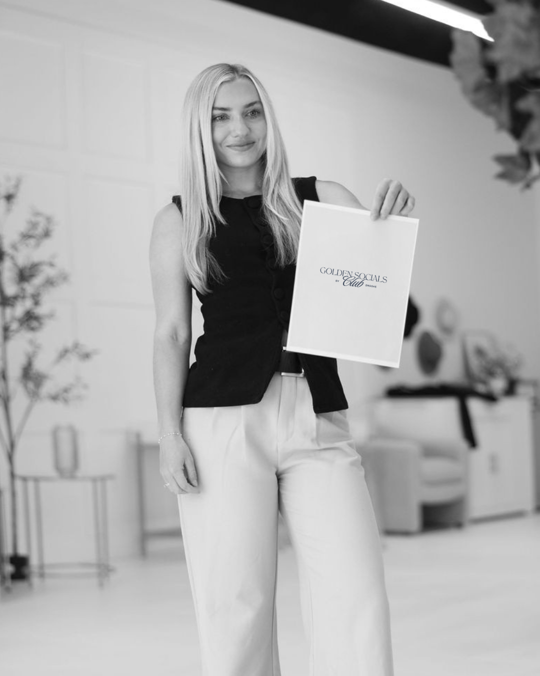 A young woman with long blonde hair smiling and holding a framed certificate or award that reads 'GOLDEN SOCIALS by Club Brooke'. She is dressed in a sleeveless black top and light-colored pants, standing indoors with a blurred background of furniture and plants.