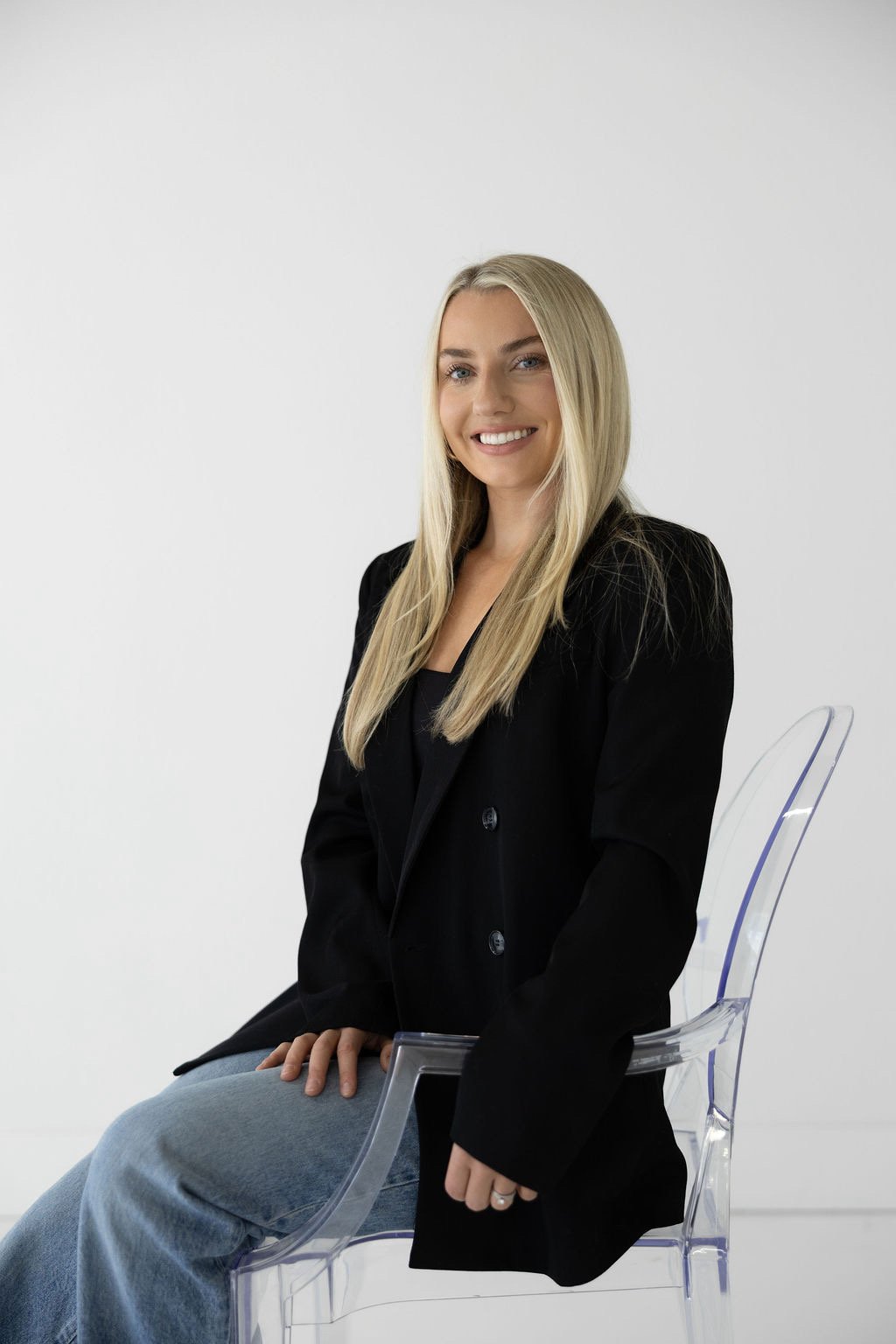 A young woman with long blonde hair, smiling, wearing a black blazer and jeans, sitting on a clear modern chair against a plain white background.