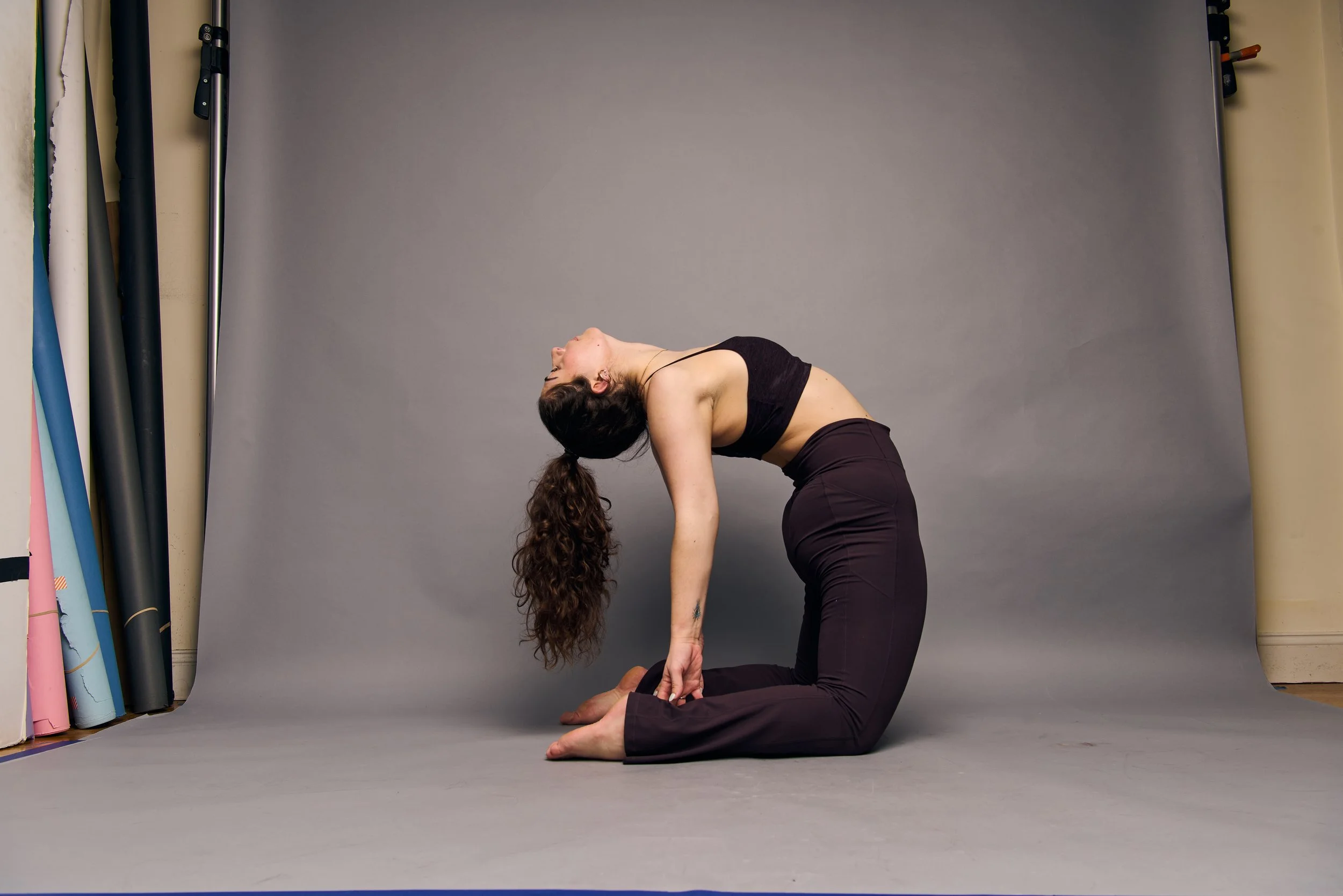 A woman practicing yoga in a studio, performing a backbend on her knees with her hands reaching toward her feet.
