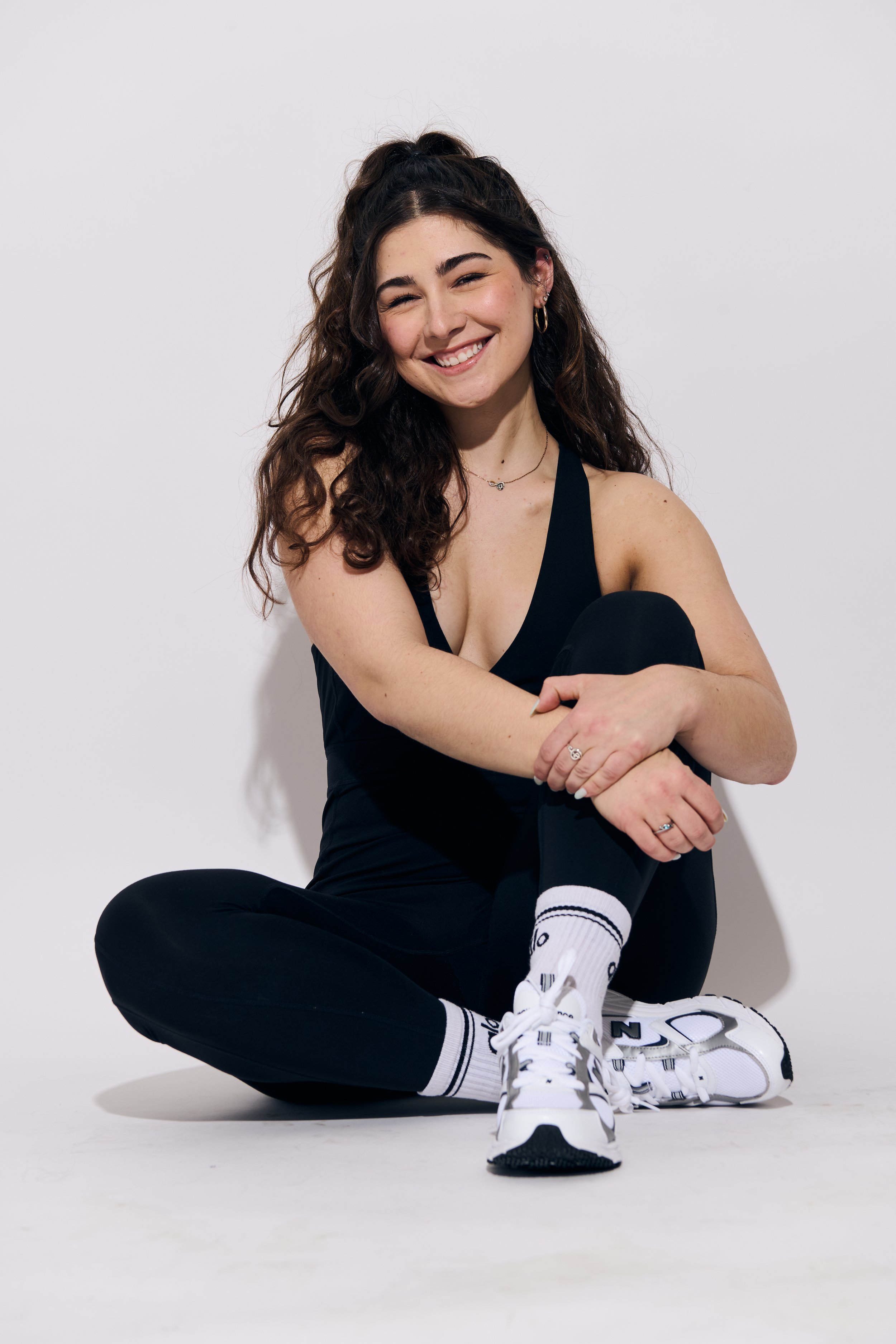 Young woman with long curly hair smiling, sitting cross-legged, wearing black tank top, black leggings, white sneakers, and white socks with black stripes, against a plain white background.