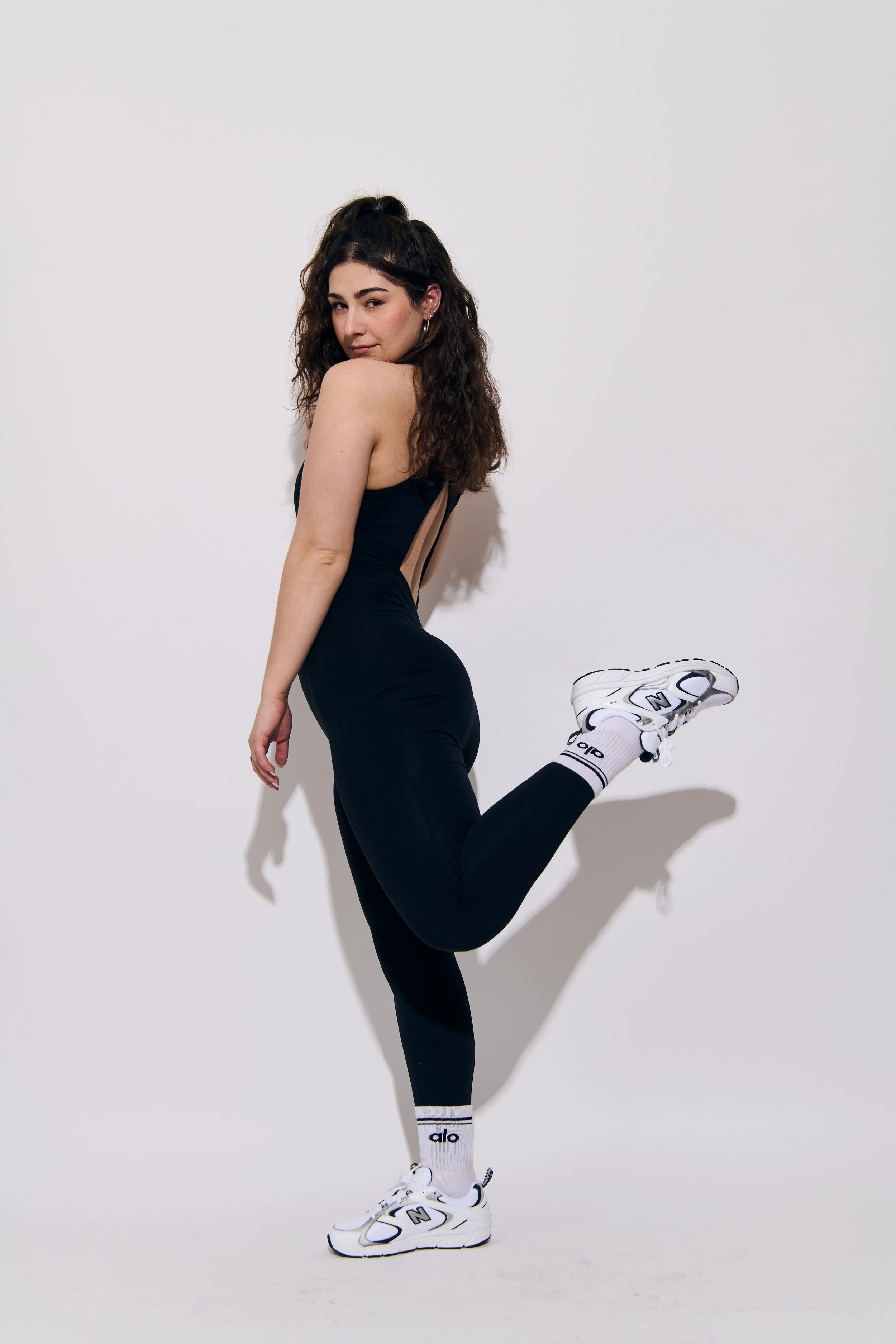 A young woman with long curly dark hair is posing against a plain white wall, wearing black athletic clothes, white sneakers, and white socks with black stripes and the word 'alo'. She is standing on one leg with her other leg bent behind her, holding her foot with one hand while looking at the camera.
