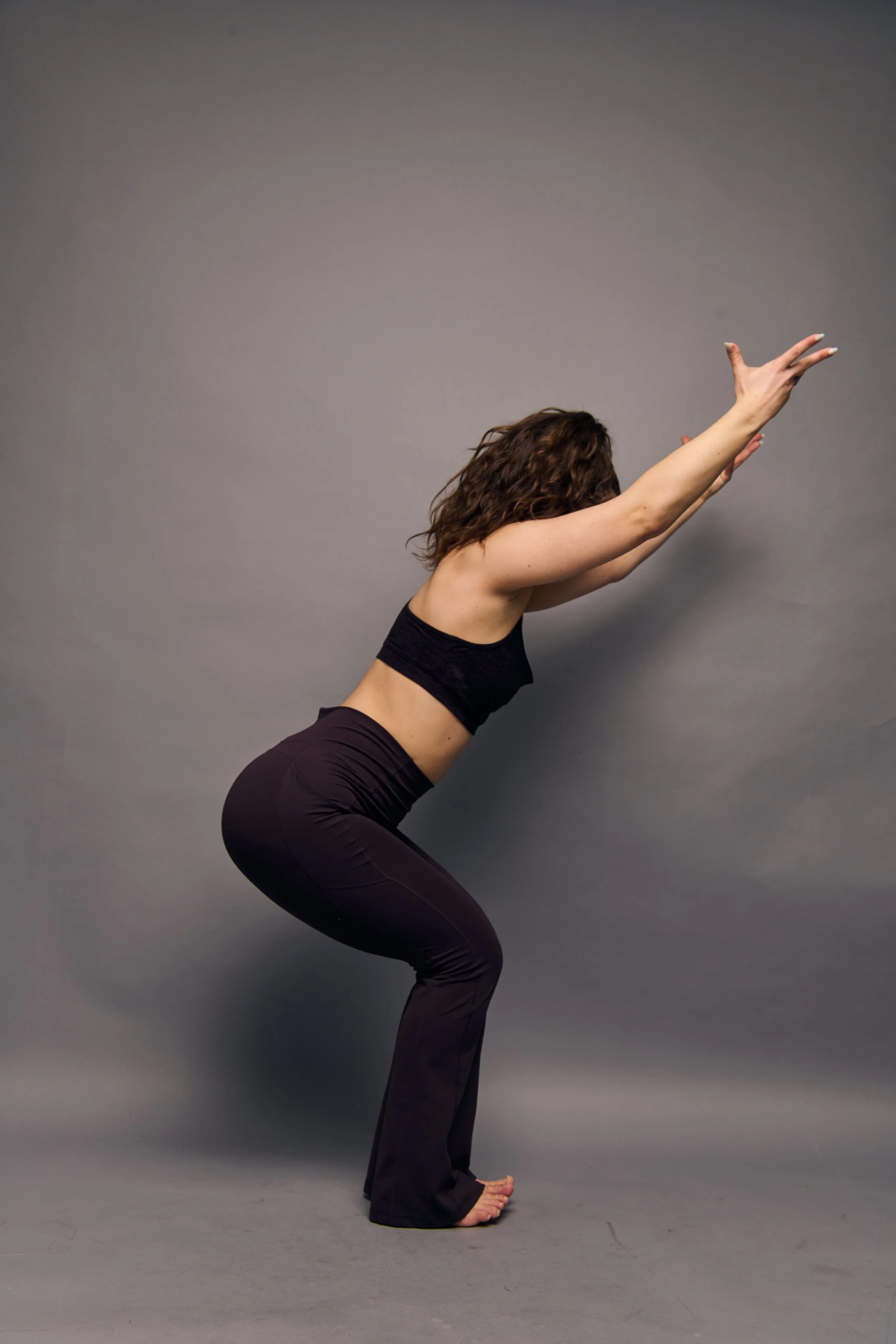 A woman with curly hair performing a yoga pose called forward bend, with her arms stretched forward and her head lowered, standing on a gray studio floor against a neutral gray background.