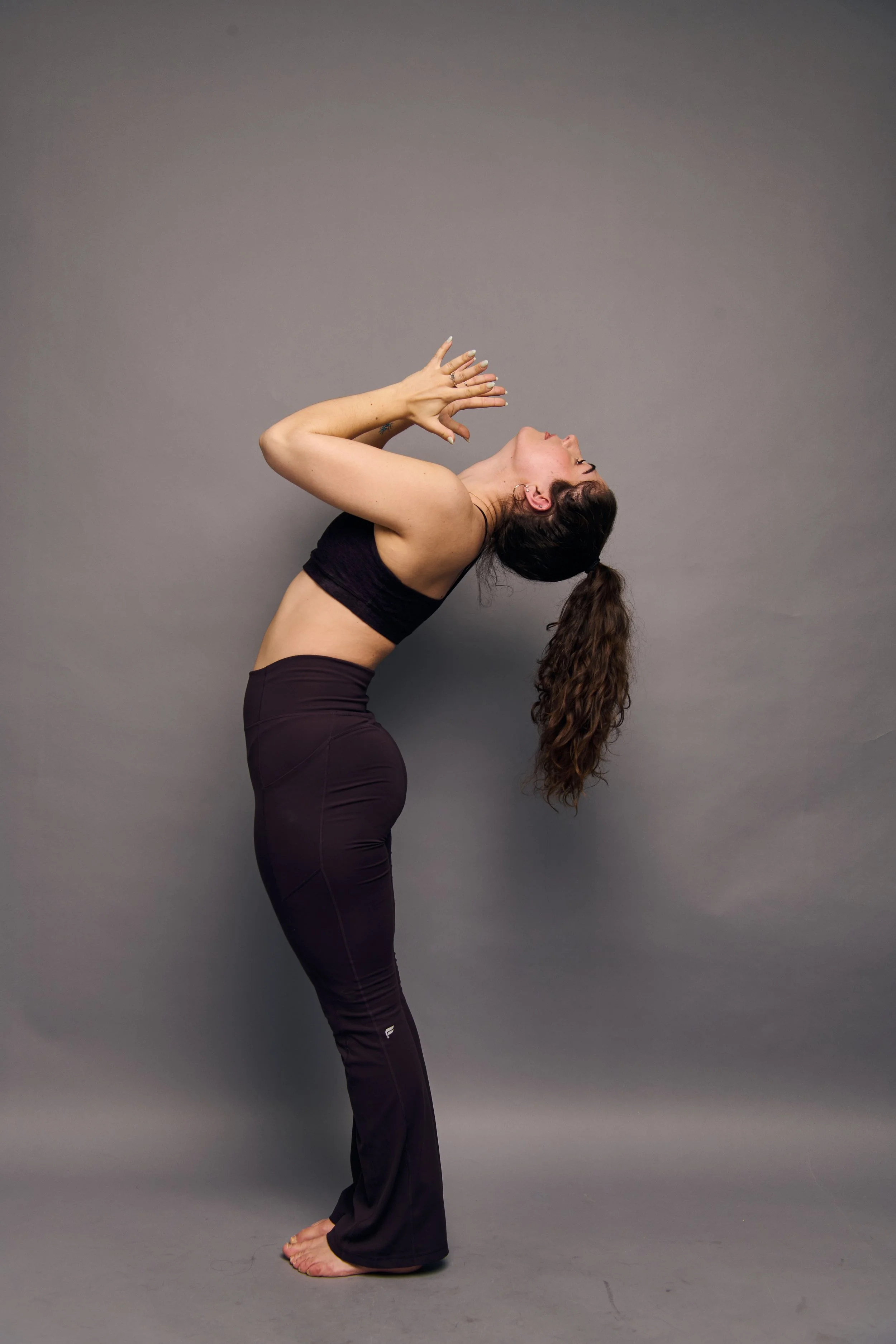 A woman practicing yoga poses against a plain gray background. She is bending backward with her hands raised near her head, fingers spread, and her long hair hanging down. She is wearing a black sports bra and matching black yoga pants.