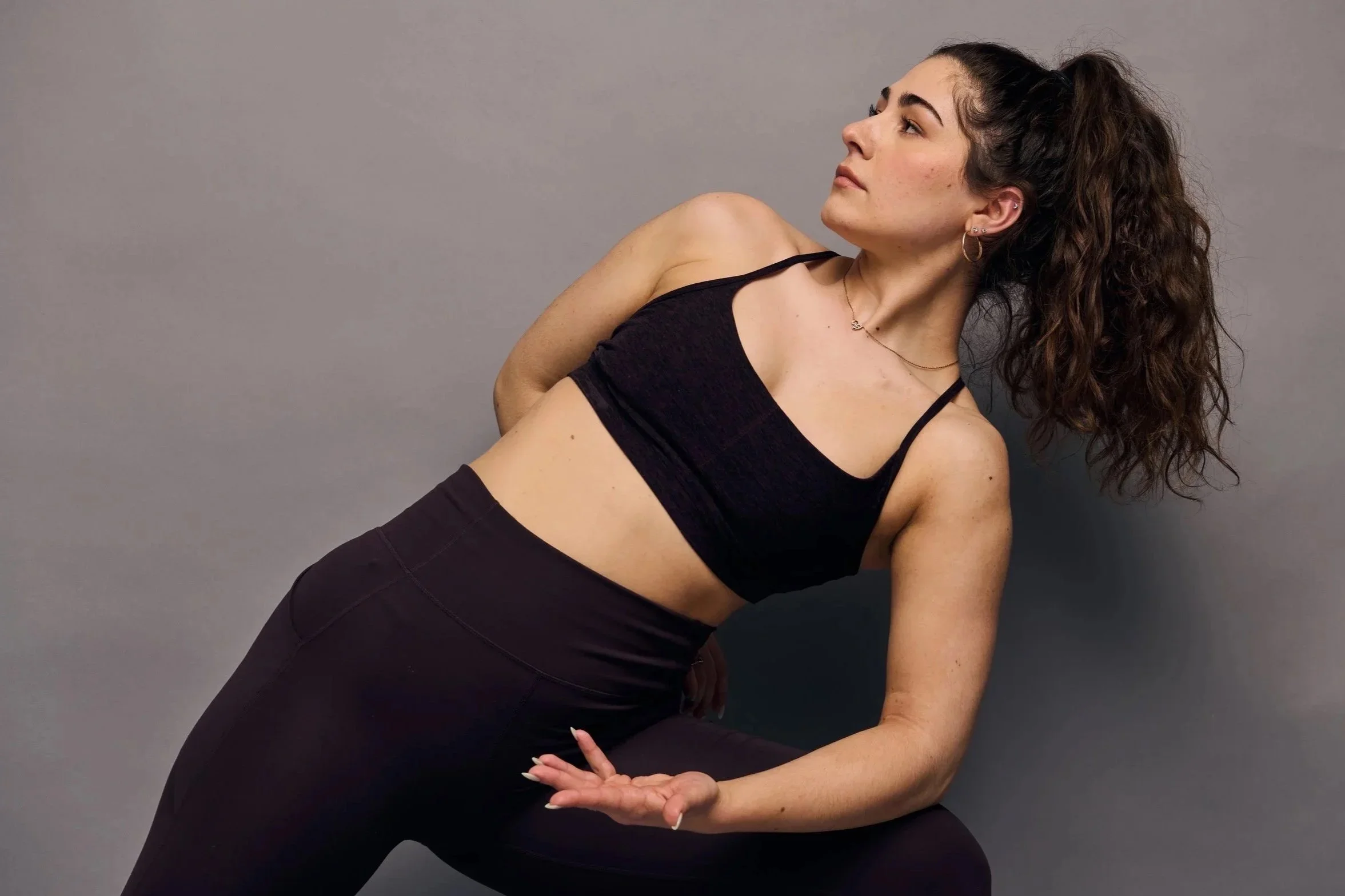 A young woman with long, curly brown hair tied in a ponytail, wearing a black sports bra and black pants, is lying on her side against a gray background, looking to the right with a relaxed expression.