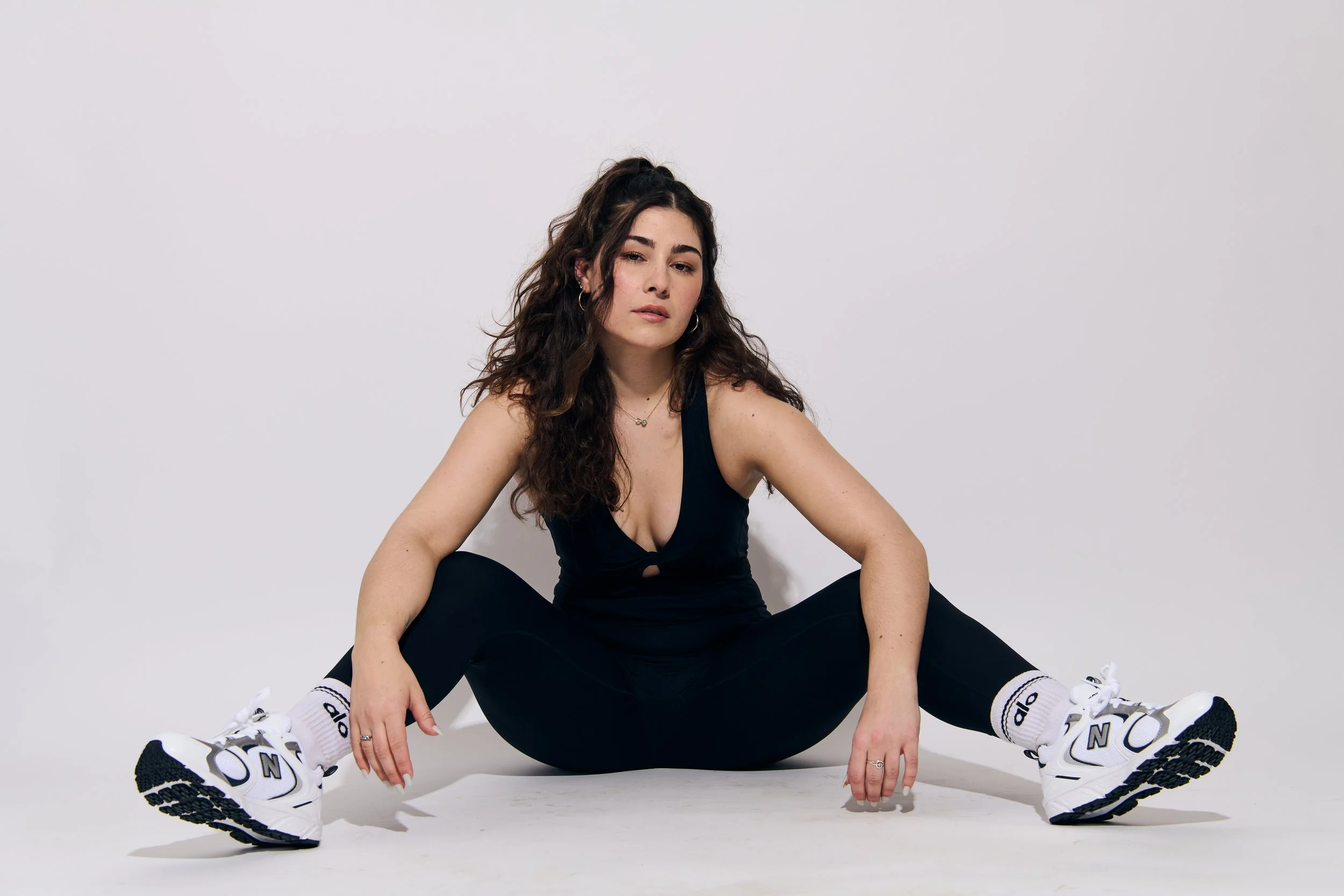 Young woman with long curly hair sitting on the floor against a plain gray background, wearing black athletic wear and white sneakers, with her legs apart and arms resting on her knees.