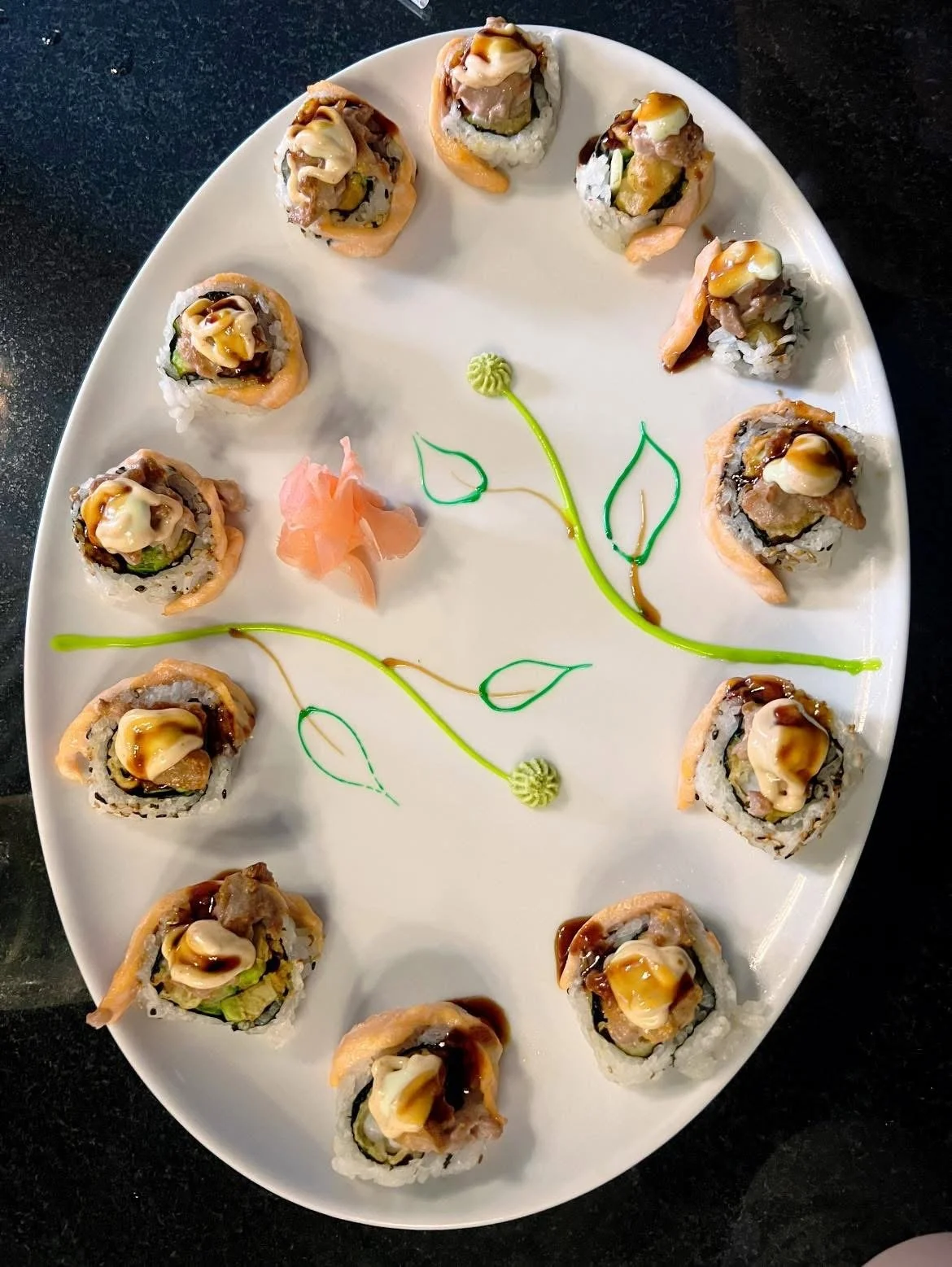 A white oval plate with 12 sushi rolls decorated with green and yellow sauce, and a small piece of ginger in the center, arranged around green icing piping in a floral pattern on a black surface.