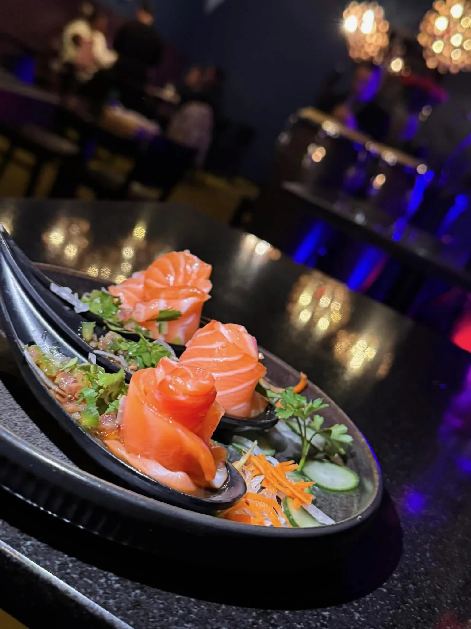 Close-up of a serving dish filled with smoked salmon, garnished with greens and vegetables, set on a dark countertop in a restaurant with blue and yellow lighting.