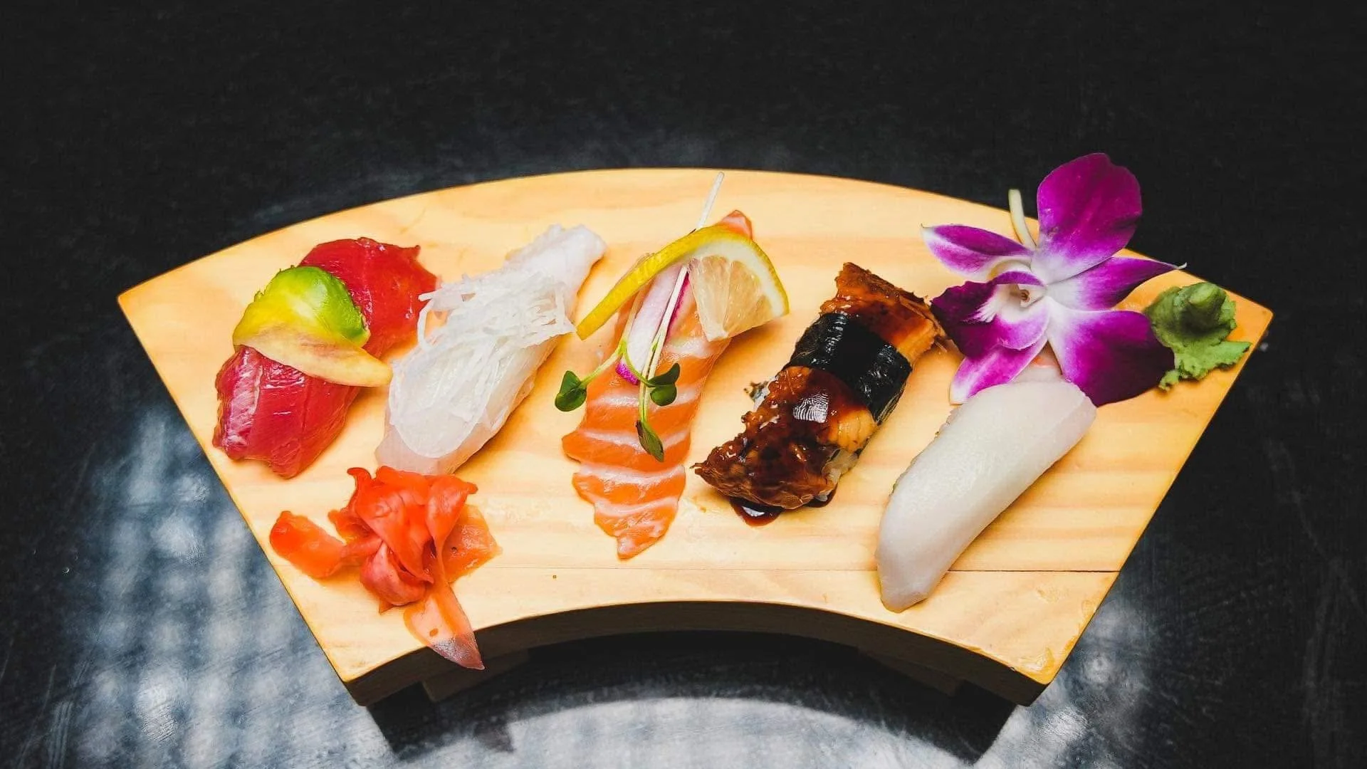 Assorted sushi pieces on a wooden platter, garnished with flower and greenery, against a dark background.
