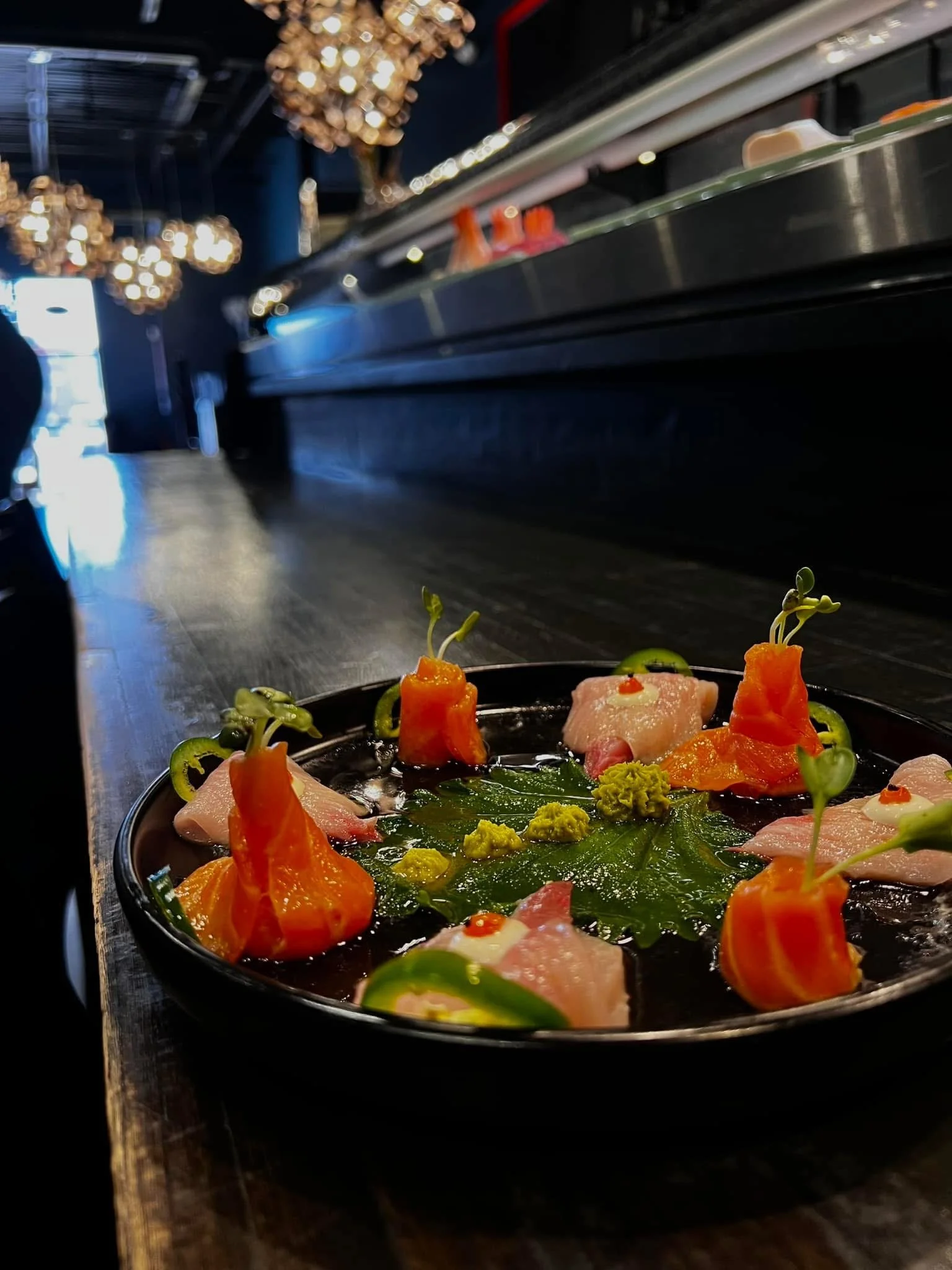 Close-up of a gourmet dish with thinly sliced fish, orange bell peppers, and green herbs, garnished with small dollops of sauce, on a black plate in a restaurant kitchen.