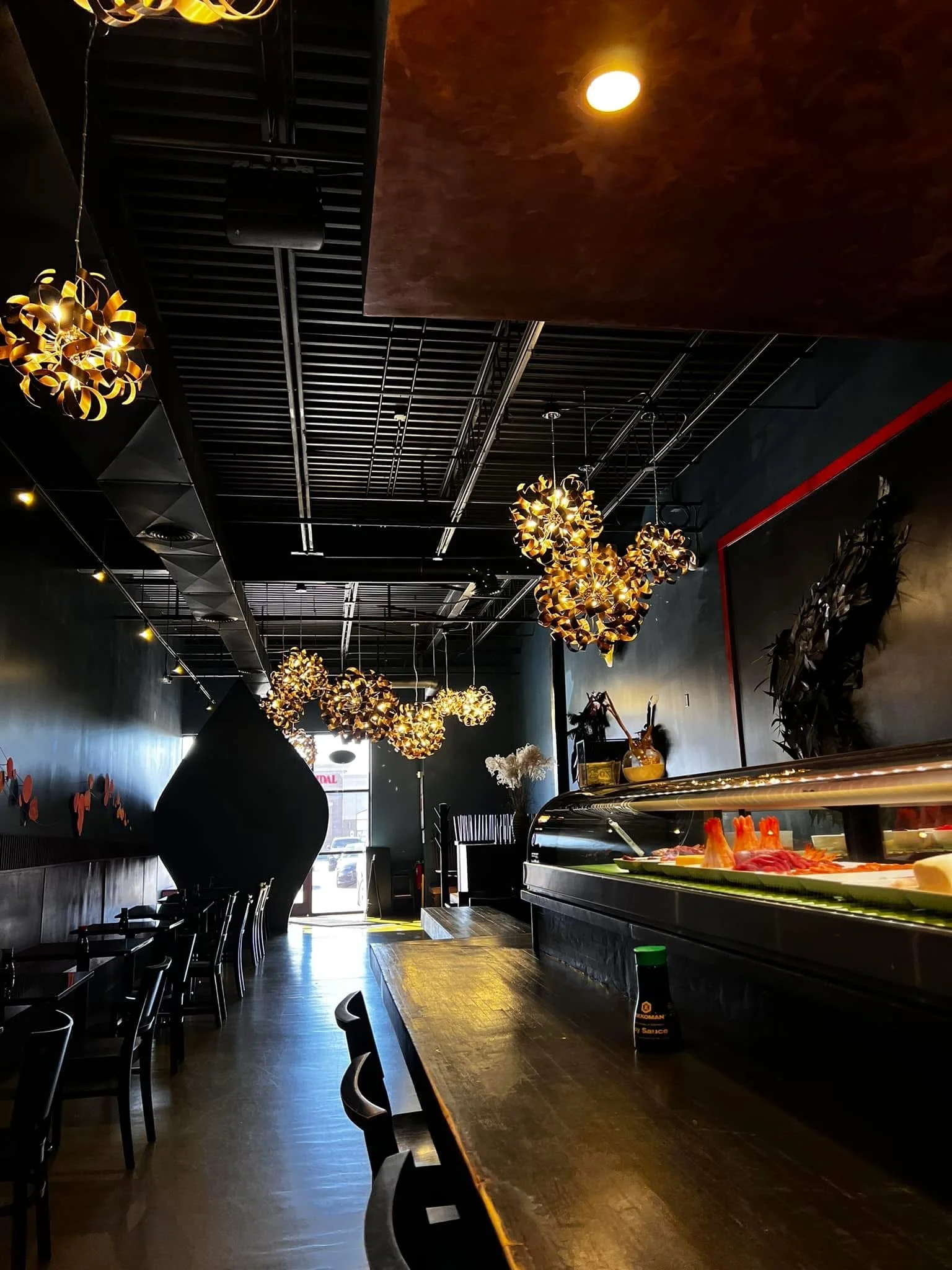 A dimly lit restaurant interior with decorative hanging lights, black chairs along a wooden counter, and a sushi display with various sashimi slices. A large black vase or sculpture is visible on the left side, and in the background, there is a doorway letting in natural light.