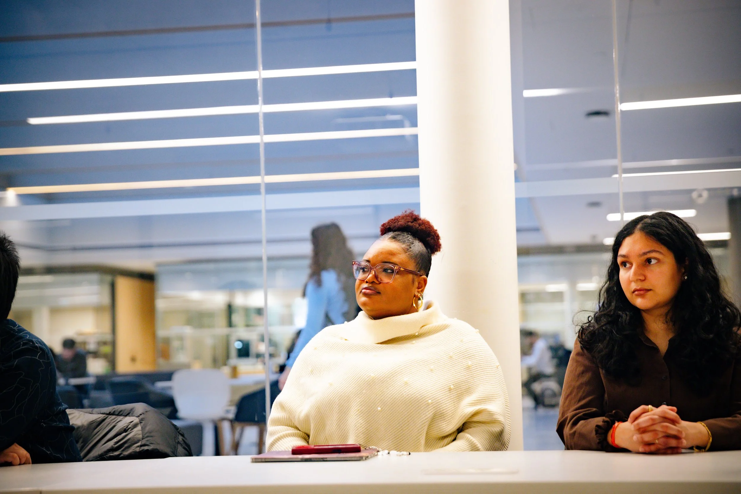 Three women sitting at a table in a modern indoor setting with ceiling lights, engaged in conversation or listening.