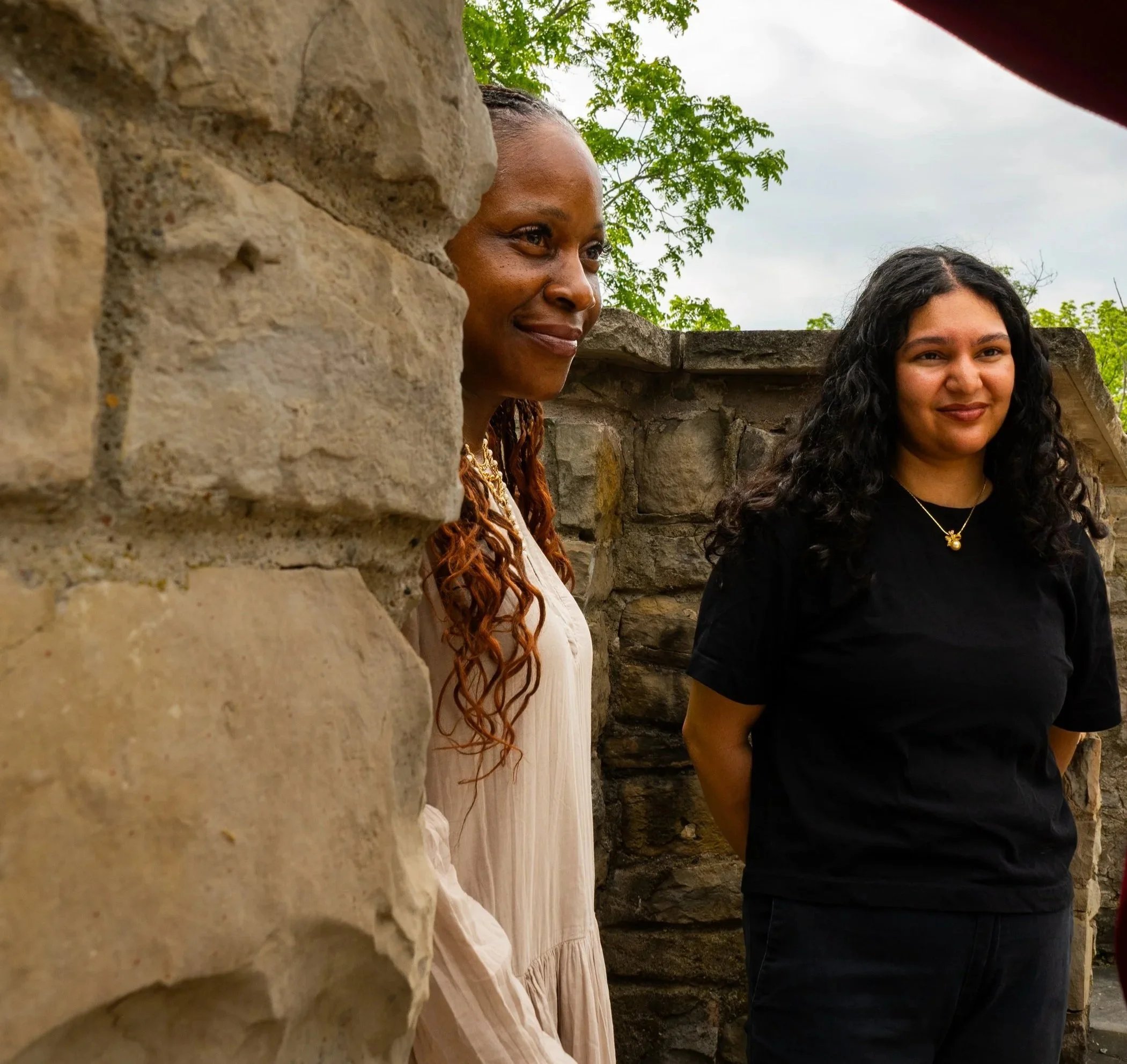 Two women standing outdoors behind a large stone or concrete structure, with greenery and a cloudy sky in the background.
