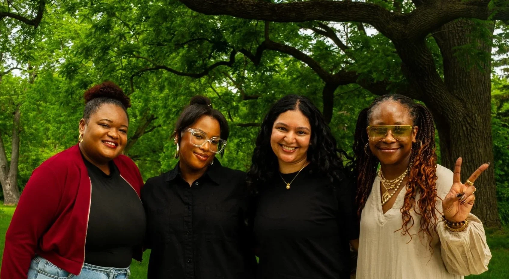 Four women standing outdoors in front of a large tree with green leaves, smiling and posing for the camera. One woman on the right is making a peace sign.