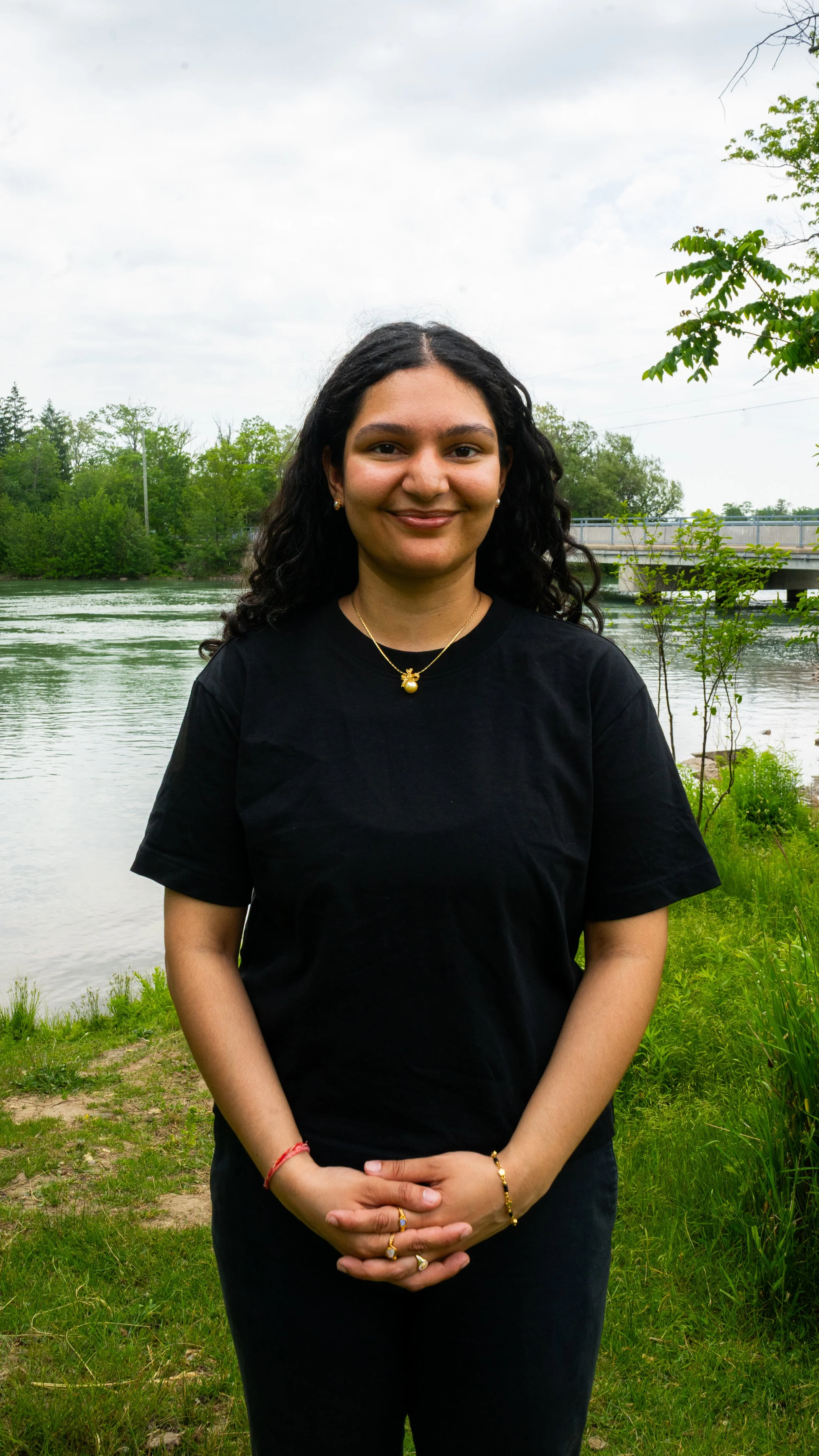 A young woman with curly black hair wearing a black t-shirt, standing outdoors by a river, with green trees and a bridge in the background.