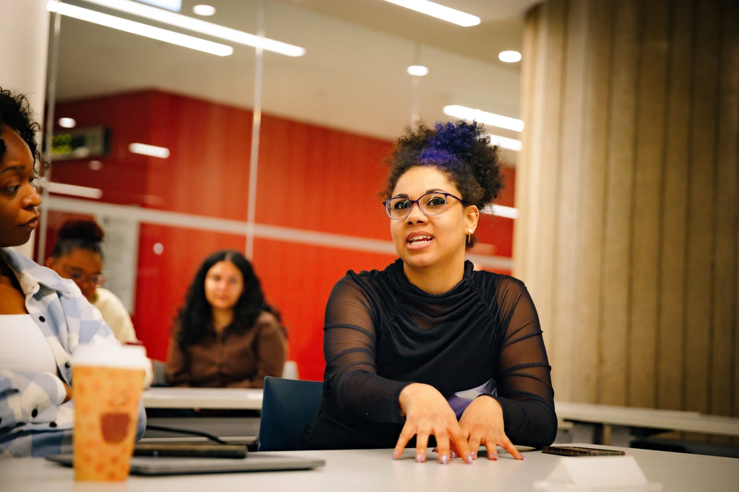 A group of diverse women sitting around a table in a classroom or meeting room, engaged in conversation. One woman in glasses and a sheer black top is gesturing with her hands while speaking. Other women are listening and watching her, with a red accent wall and beige curtains in the background.