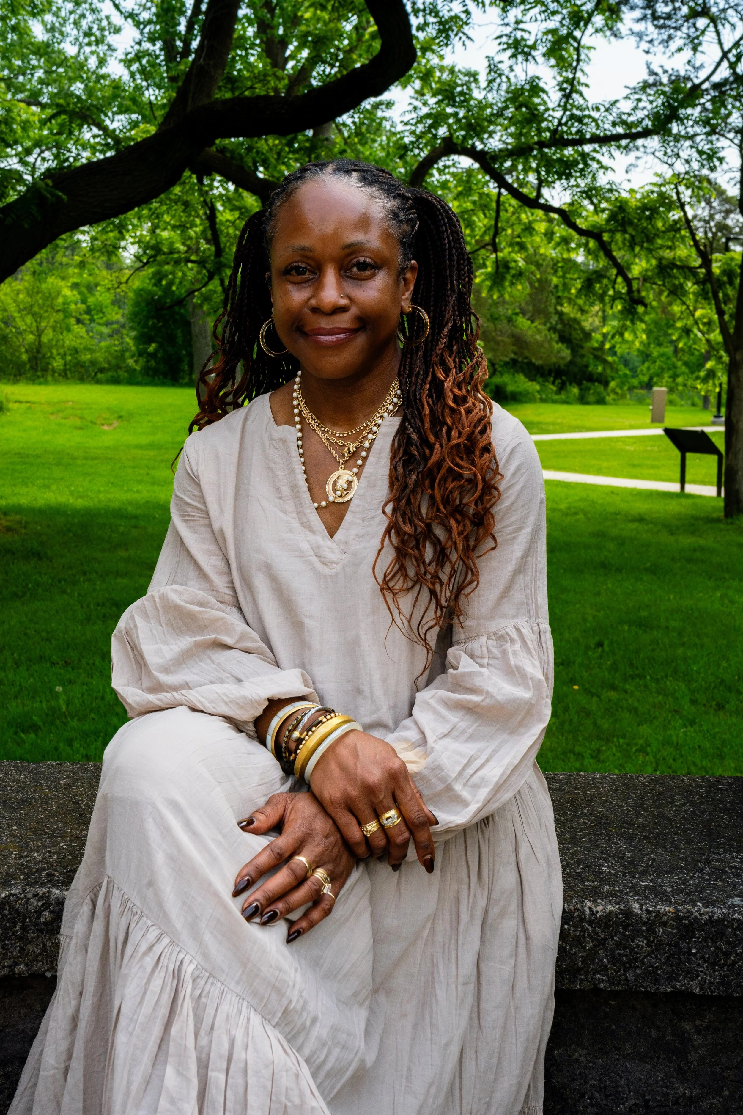 A woman sitting on a stone bench in a park with green grass and trees behind her. She has long braided hair, multiple jewelry pieces including necklaces, earrings, and bracelets, and is wearing a cream-colored dress and rings. She is smiling slightly while looking at the camera.