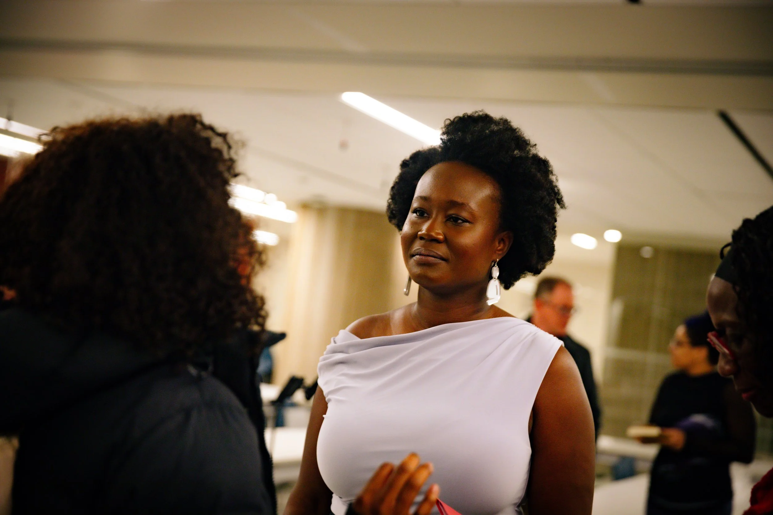 A woman with short, curly black hair wearing pearl earrings and a white off-shoulder top, engaged in conversation with two other women in a social setting.
