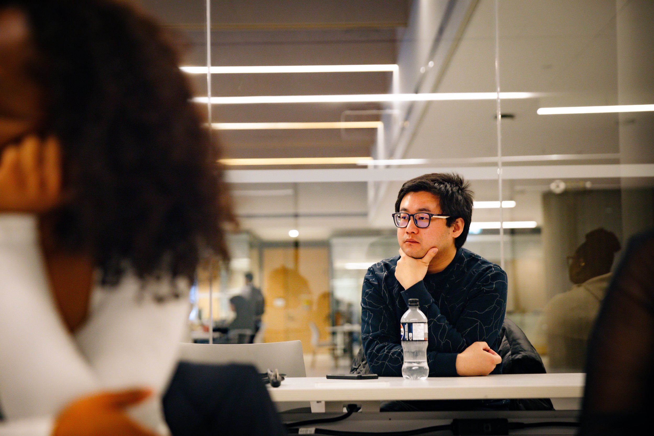 A man in glasses and a striped shirt sitting at a table in a modern, well-lit indoor space, with people blurred in the foreground.