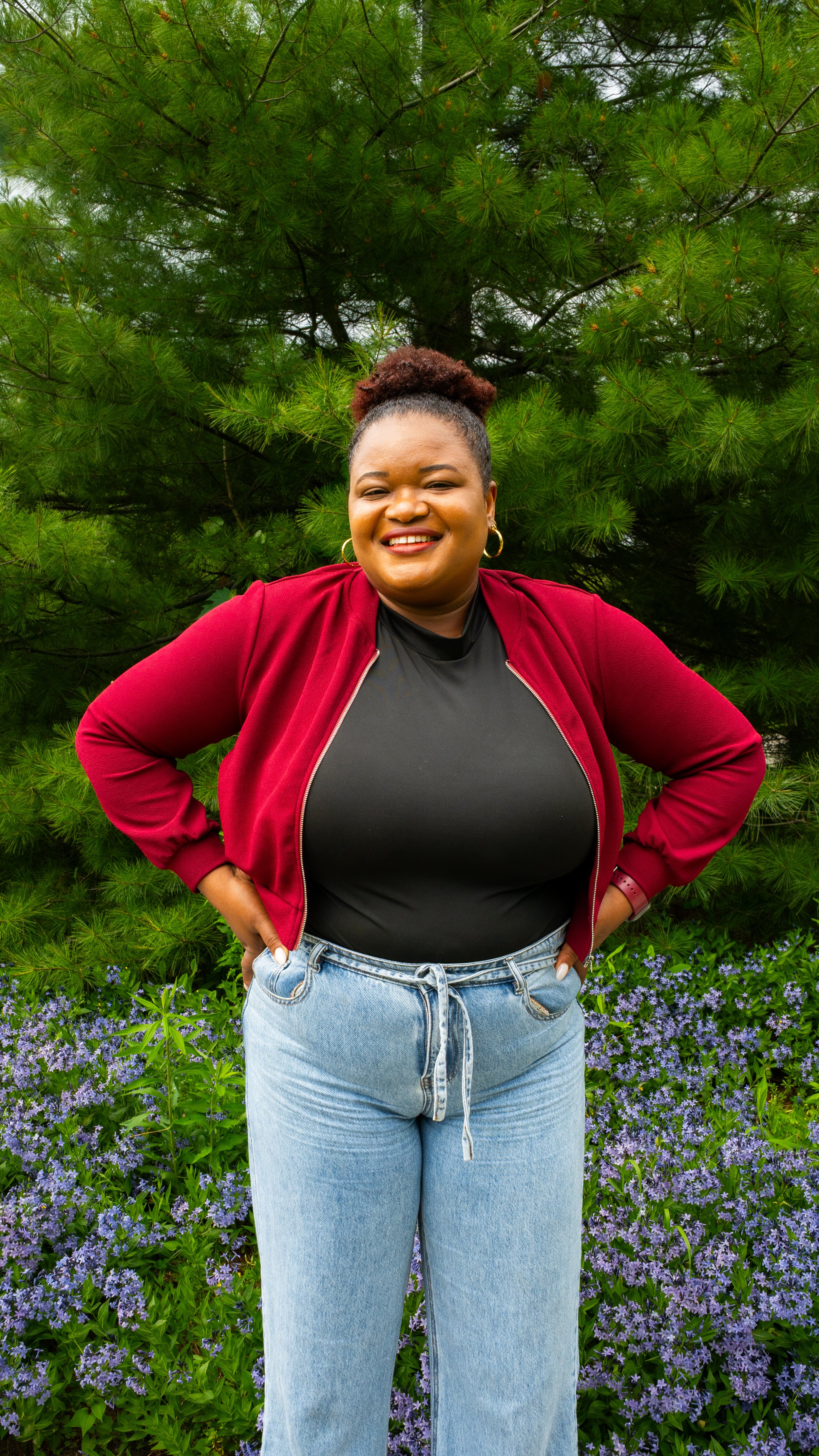 A woman standing outdoors in front of a green pine tree, smiling, wearing a red jacket, black top, and light blue jeans.
