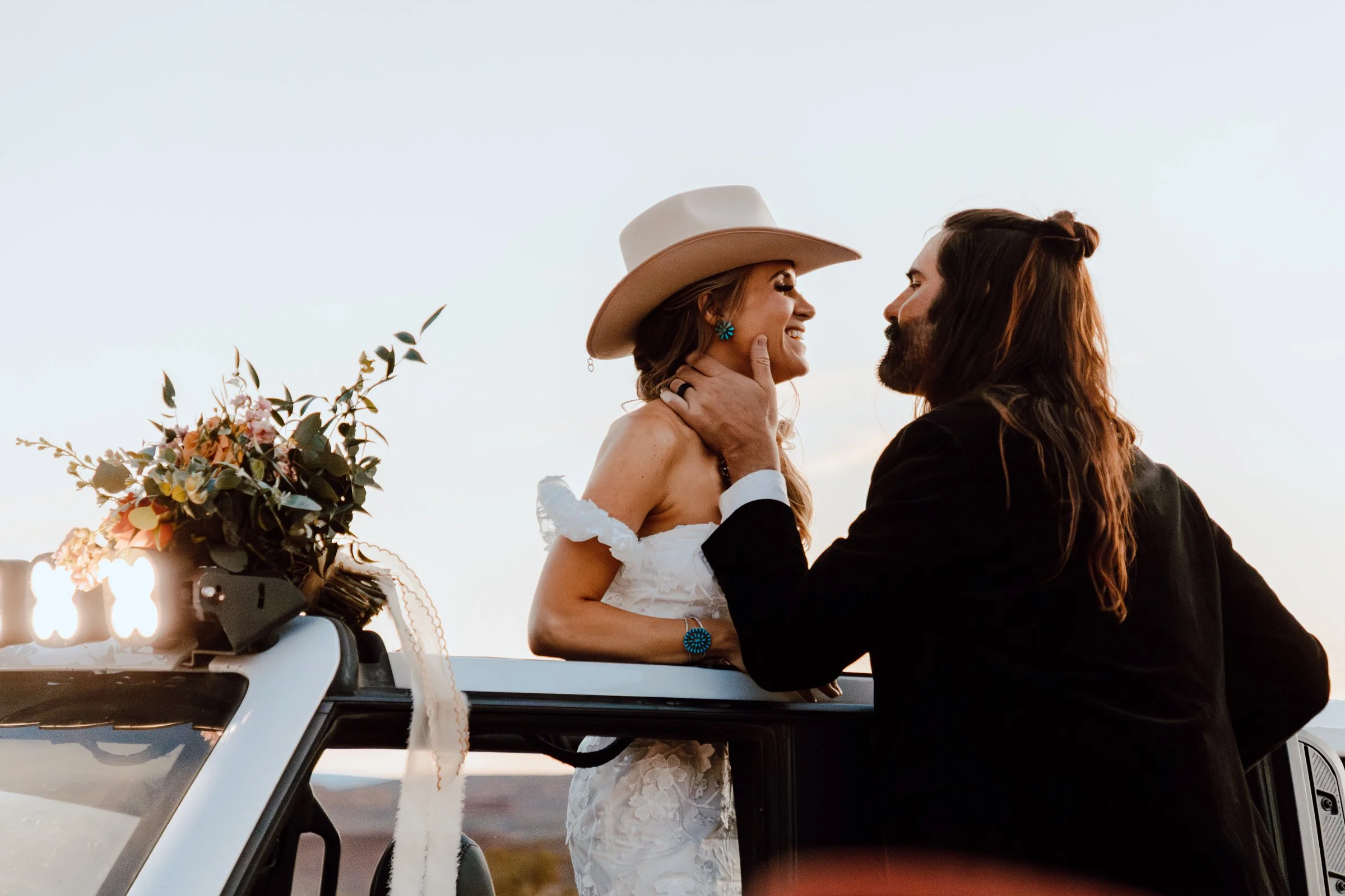 A couple in wedding attire sharing a moment on a convertible car at sunset, with the woman wearing a white dress and a cowboy hat, and the man in a black suit, with flowers on the car's hood.