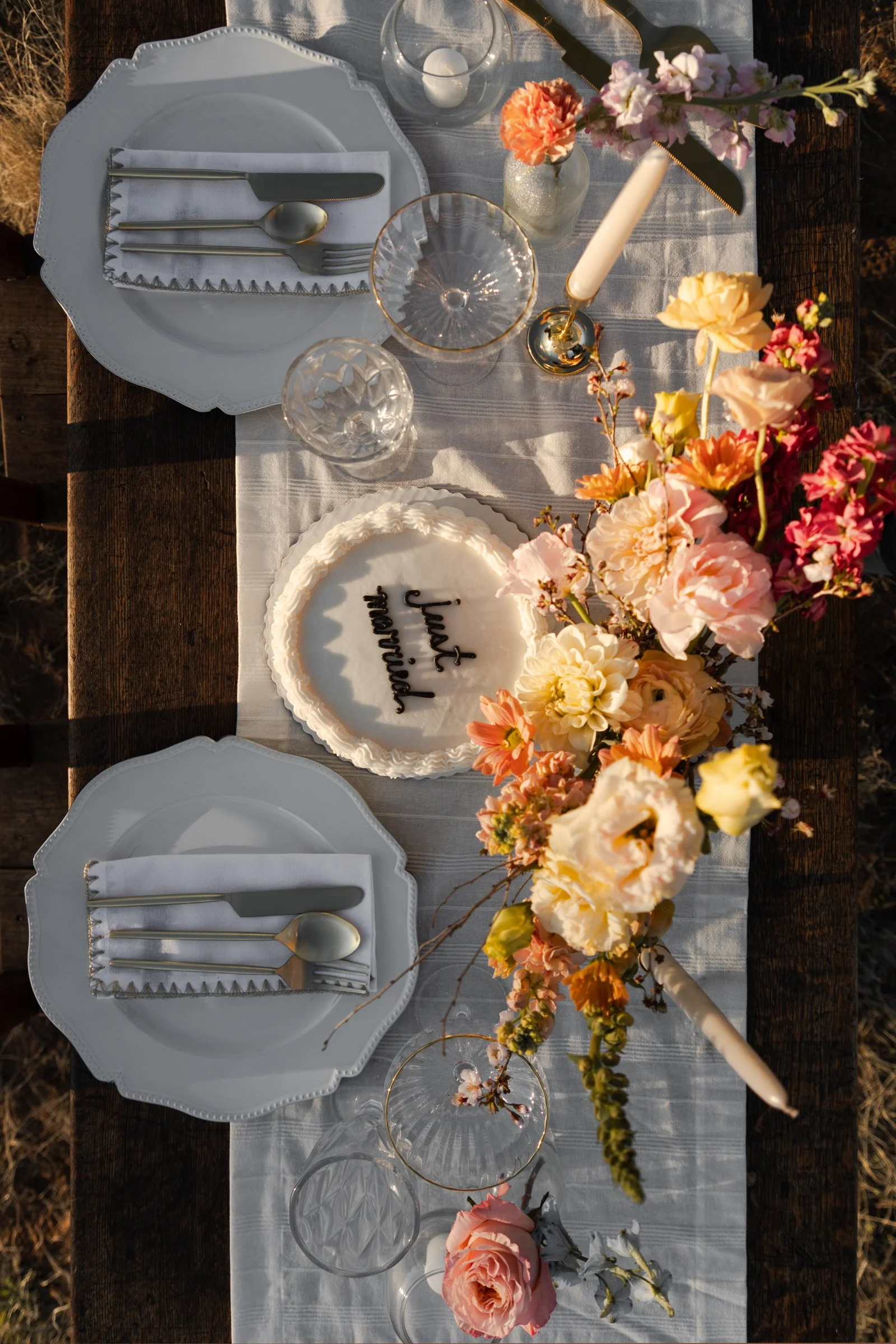 A rustic outdoor table set for a celebration with white plates, silverware, and glassware. A white cake with black writing, partially visible, is on the table. There are colorful flower arrangements and a gold-colored candle holder with a white candl