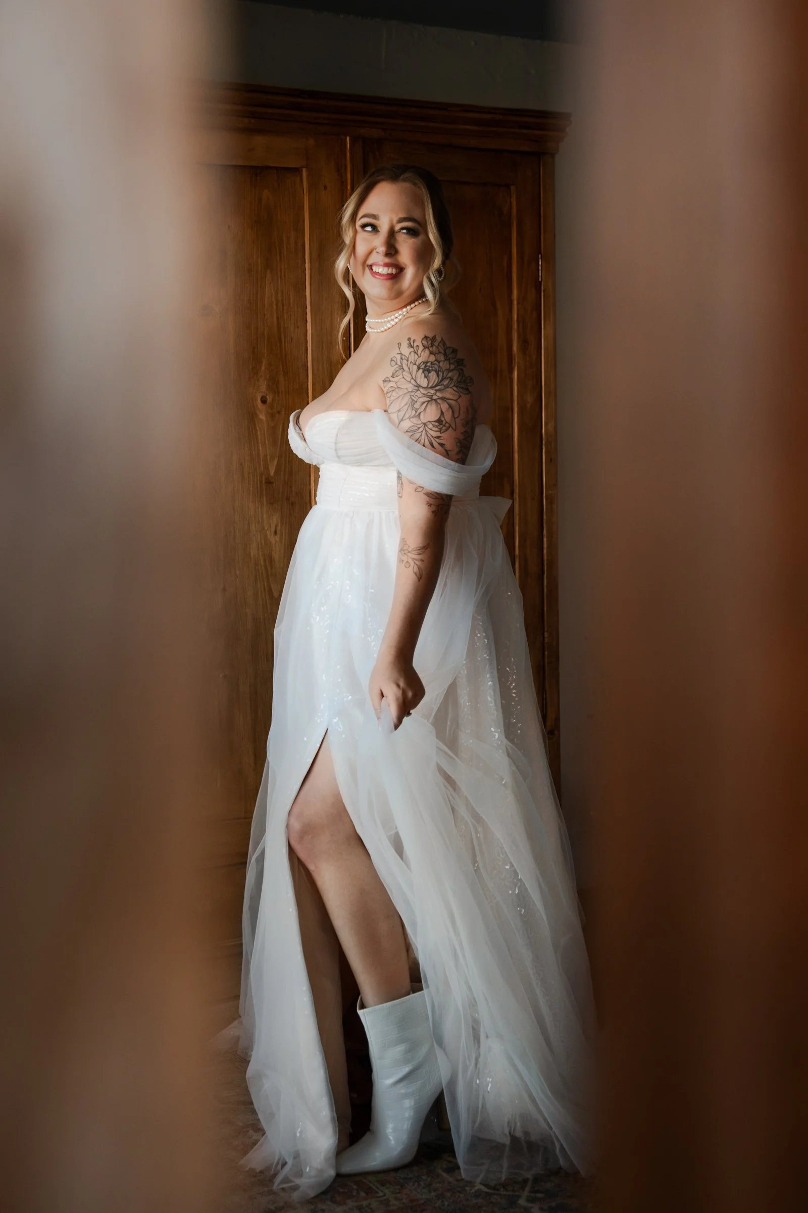 A woman in a white wedding dress with off-the-shoulder sleeves and a slit, standing in front of a wooden door, smiling and looking to the side. She has blonde hair styled in waves, wearing pearl jewelry, and has a large floral tattoo on her upper arm