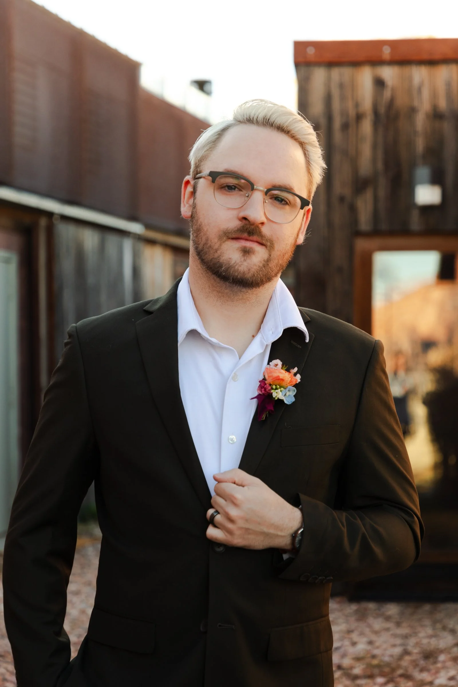 A man with glasses and a beard dressed in a black suit and white shirt, standing outdoors with wooden buildings in the background.