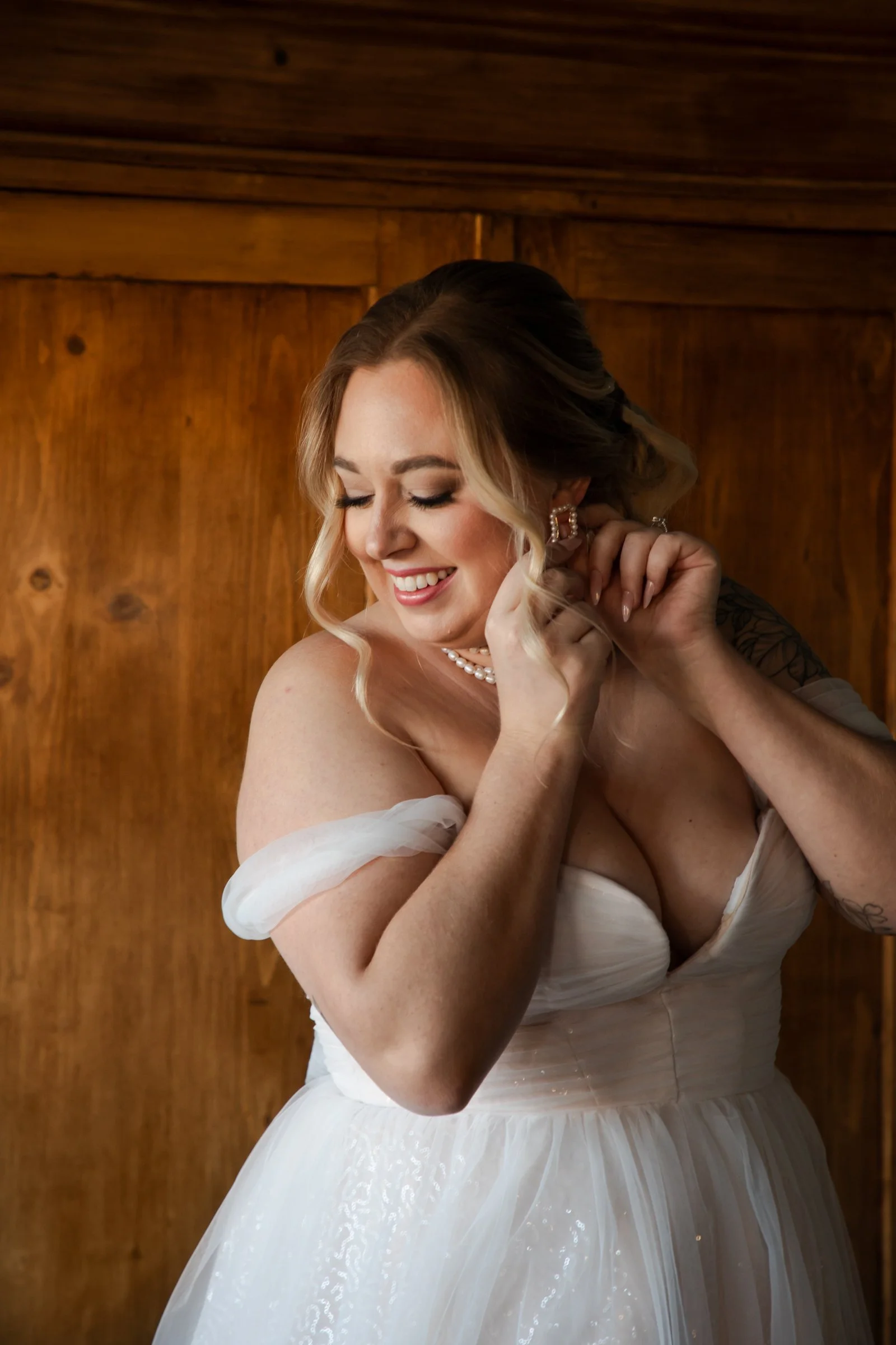 A smiling woman in a wedding dress adjusting her earring in front of a wooden wall.