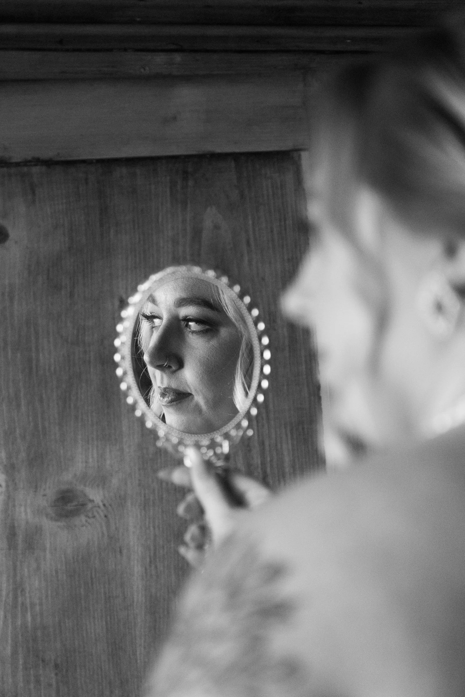 A woman looking at her reflection in a small, round mirror with a beaded edge. The mirror reflects her face, showing her eyebrows, eyes, nose, and lips. She has light-colored hair and a nose piercing. The background features a wooden wall.