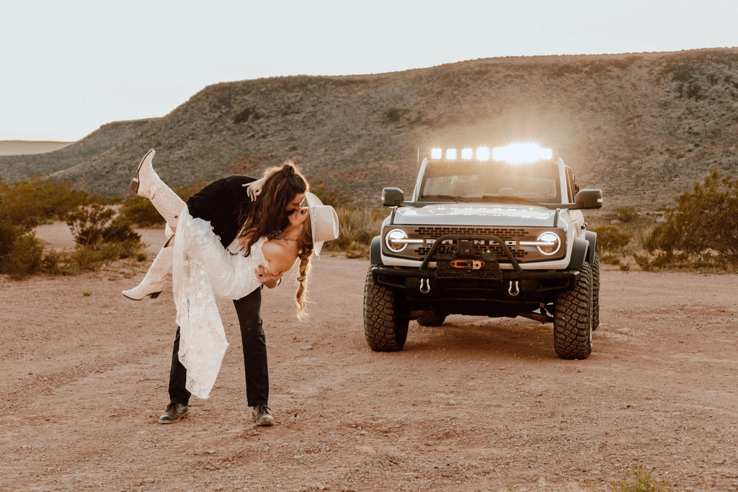 A woman wearing a white dress and cowboy boots is being dipped backward in a playful dance move in front of a gray off-road vehicle with a desert landscape and hills in the background.