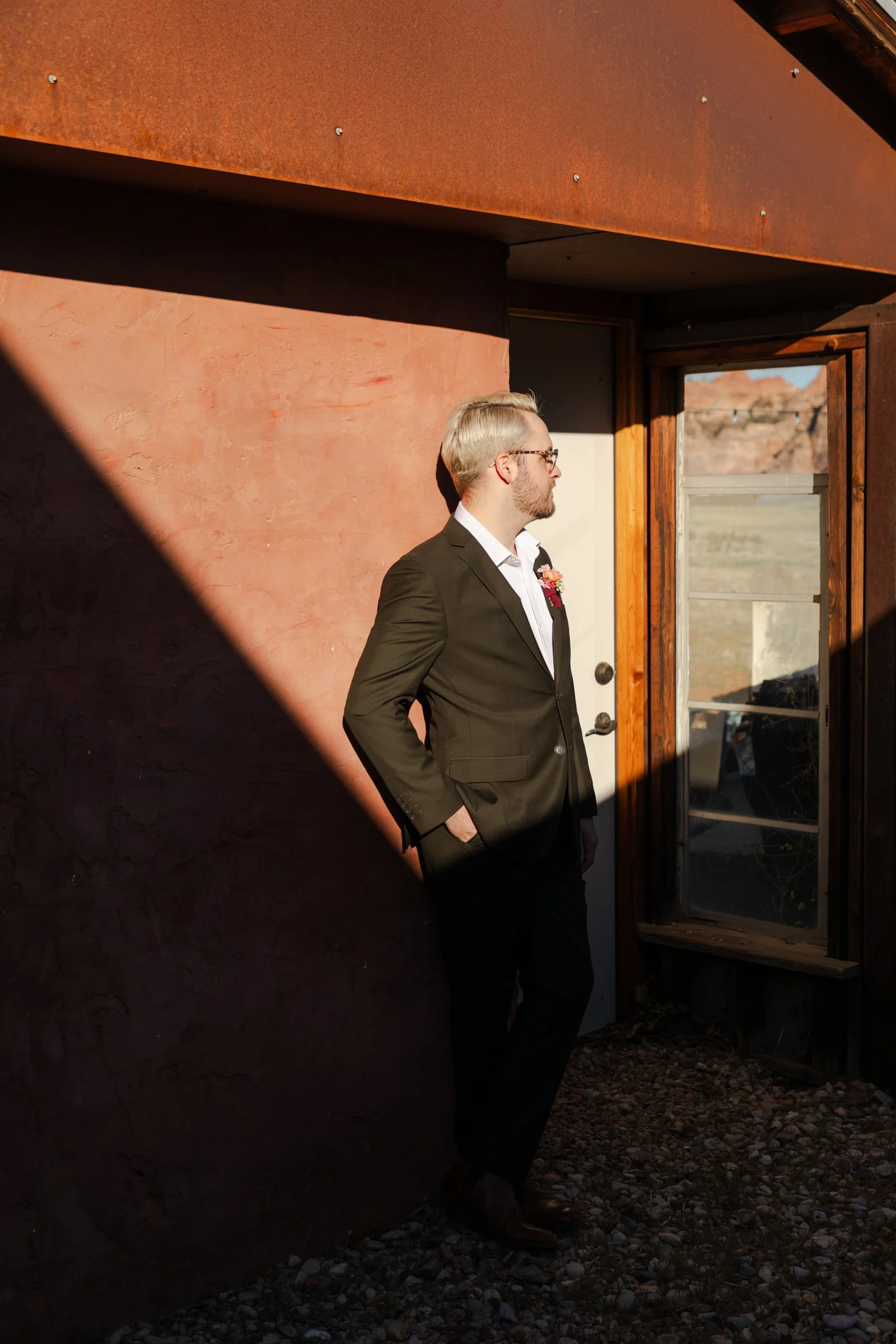 A man with blonde hair and glasses, wearing a suit, stands outside near a doorway with a scenic landscape in the background.
