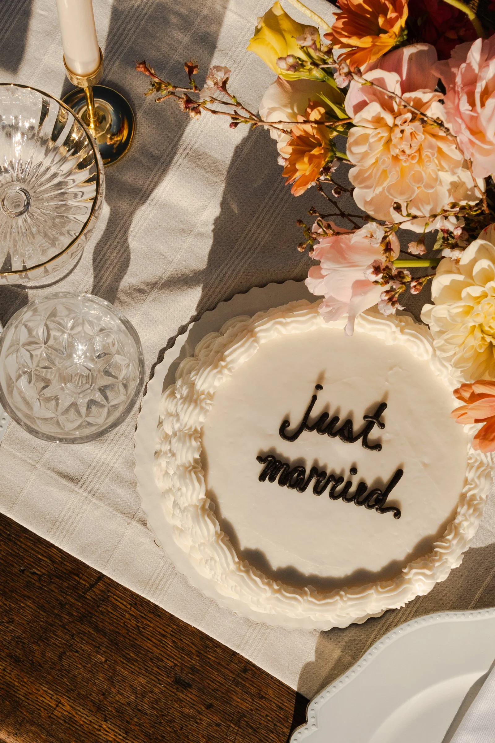 A white birthday cake with the words 'just married' written on it in black icing, surrounded by pink, orange, and cream-colored flowers, on a table with a white tablecloth. There are glassware, including a crystal bowl and a candlestick.