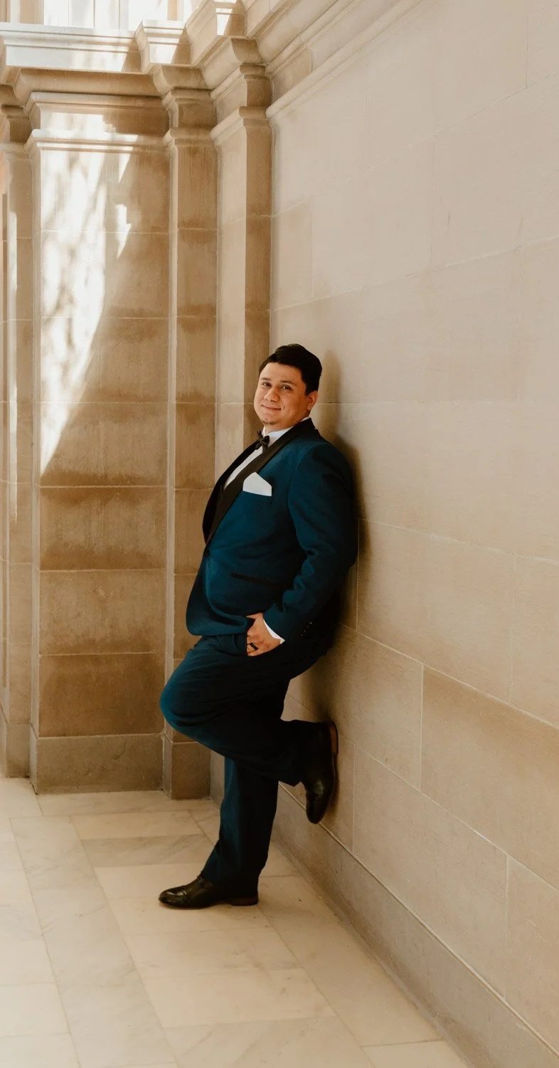 A man in a formal dark teal tuxedo with black lapel and a white pocket square, leaning against a beige stone wall inside a grand building, with one foot resting against the wall and hands in pocket, smiling at the camera.