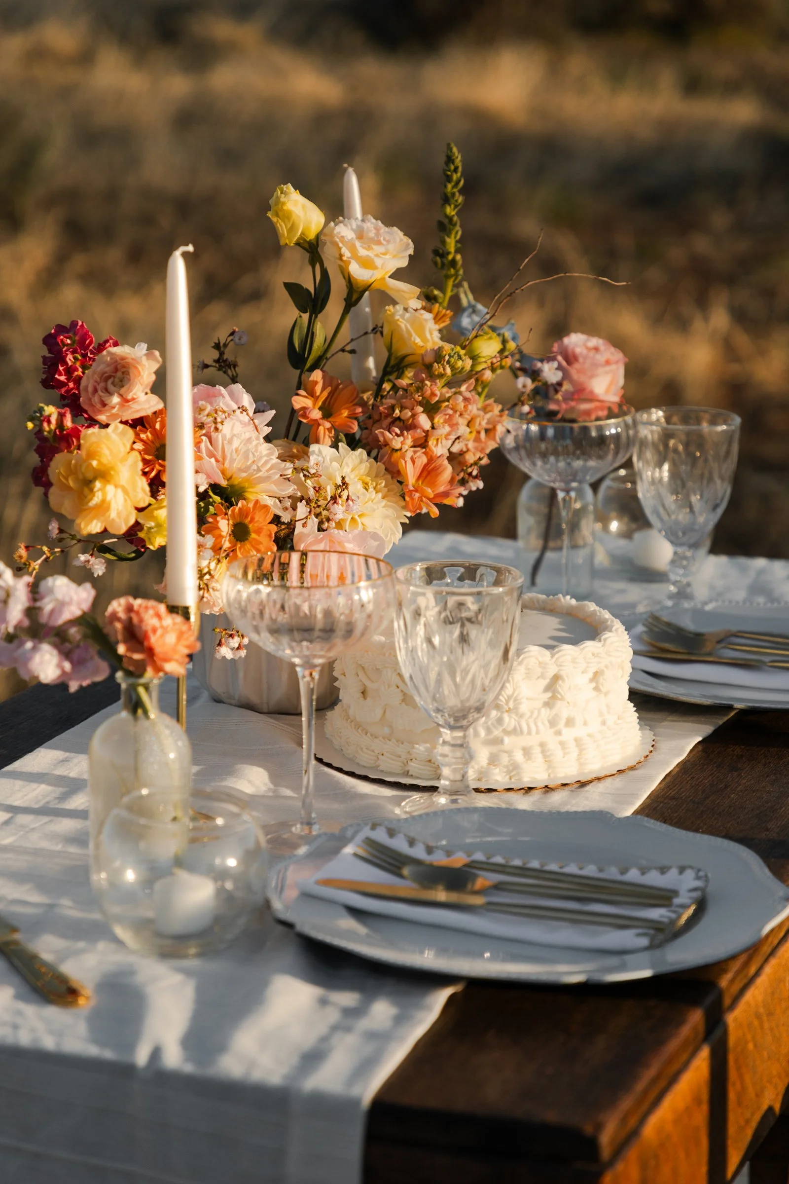 A table set outdoors with a white tablecloth, featuring a floral centerpiece with various pink, yellow, and cream flowers, white candles, glassware, plates, and gold utensils, during sunset.