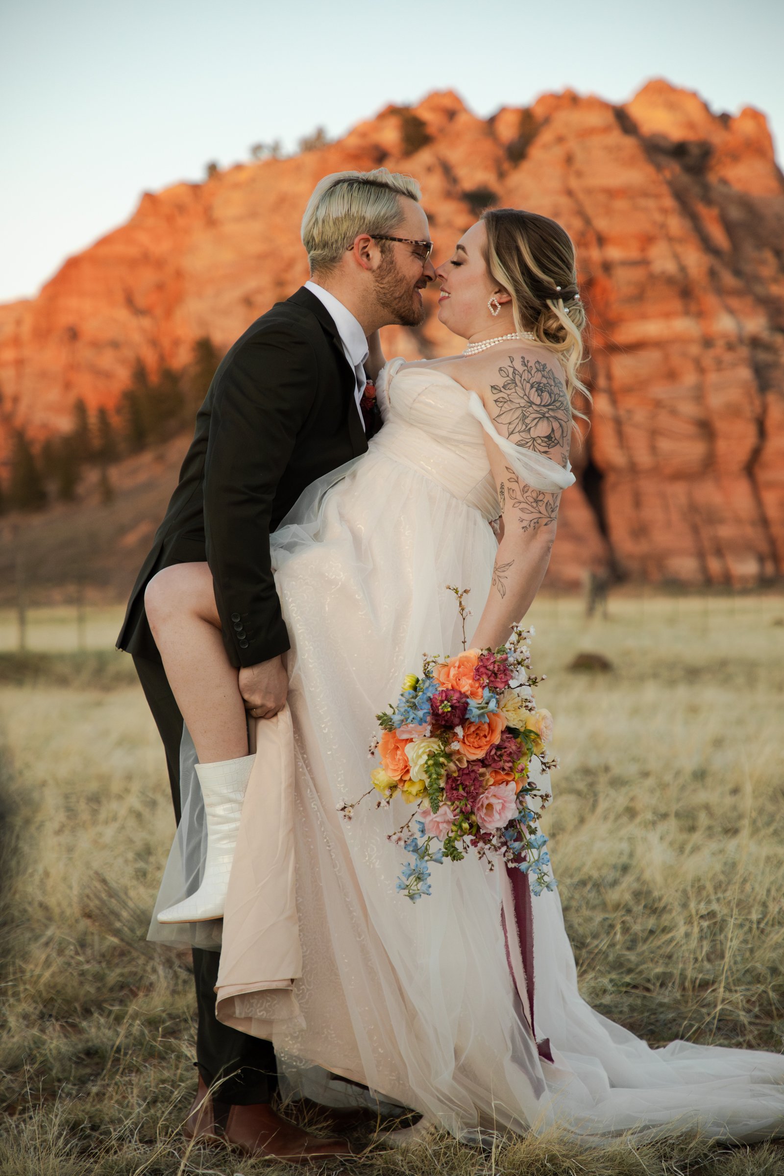 A couple in wedding attire kissing in a field with large red rock formations in the background.