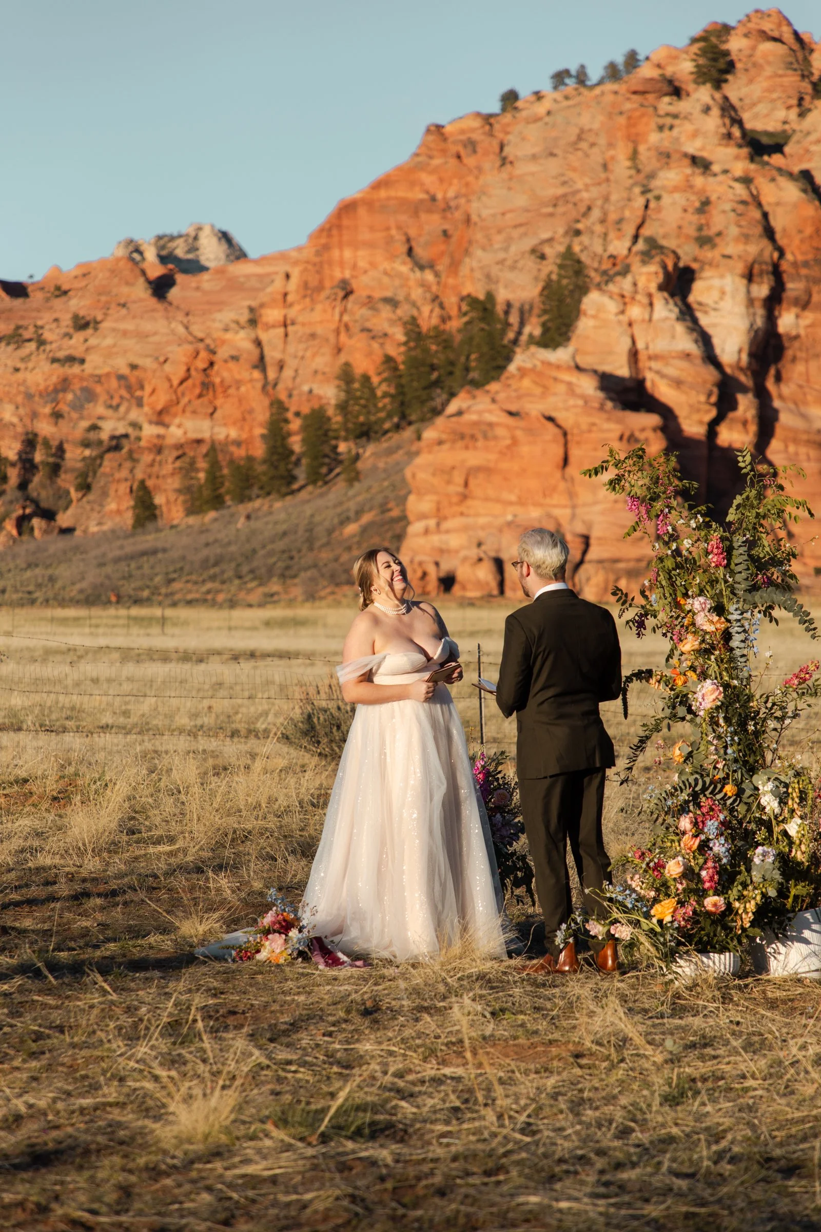 A couple getting married outdoors with red rock formations in the background, surrounded by flowers and greenery.