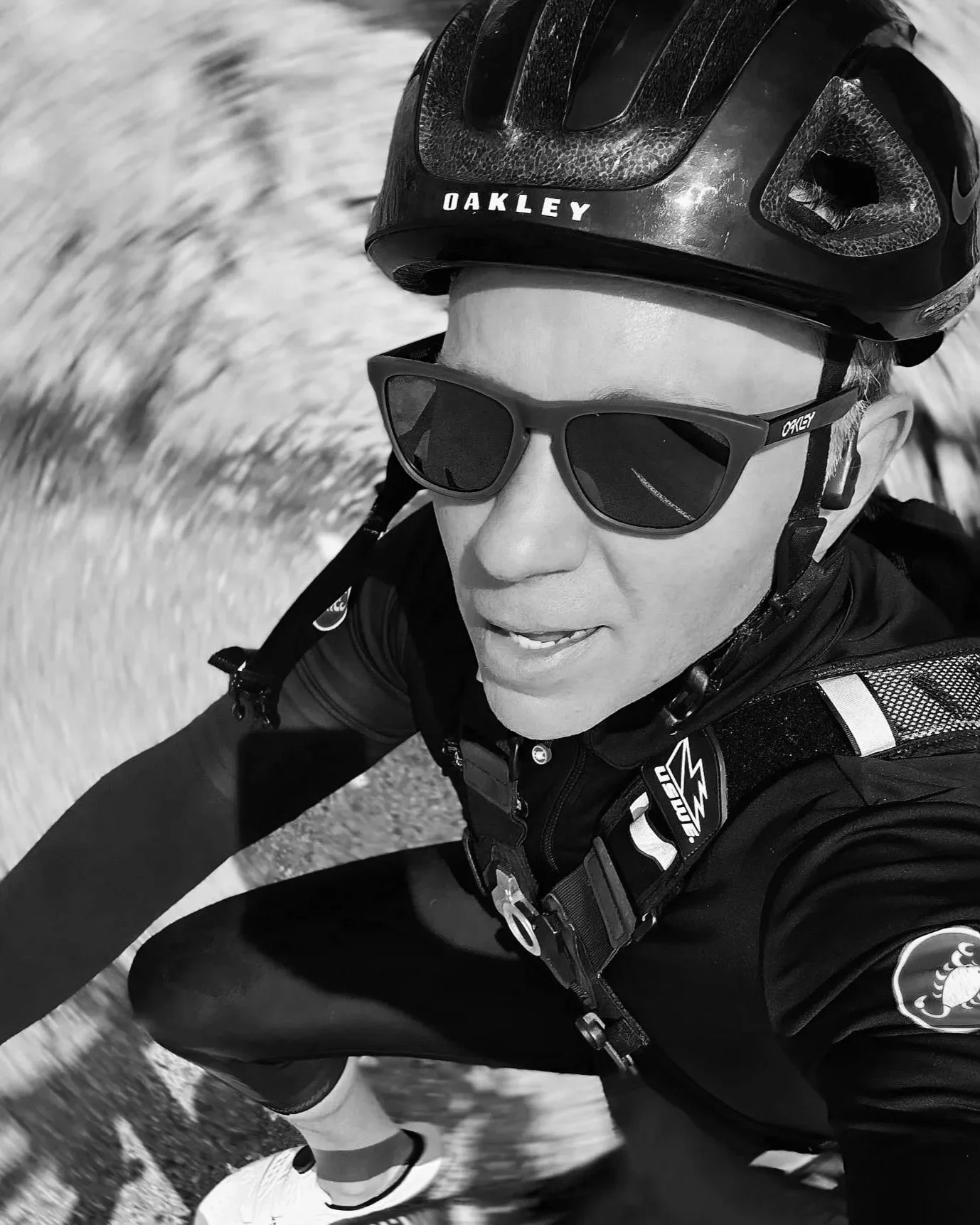 A person riding a bicycle wearing a helmet and sunglasses, captured in motion. The image is in black and white.