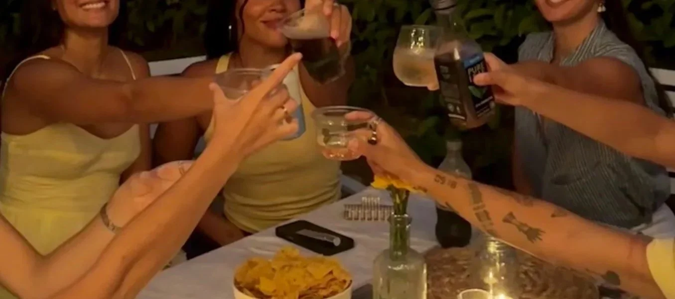 Group of friends celebrating with drinks at a dinner table, raising glasses in a toast, and a bowl of chips on the table.