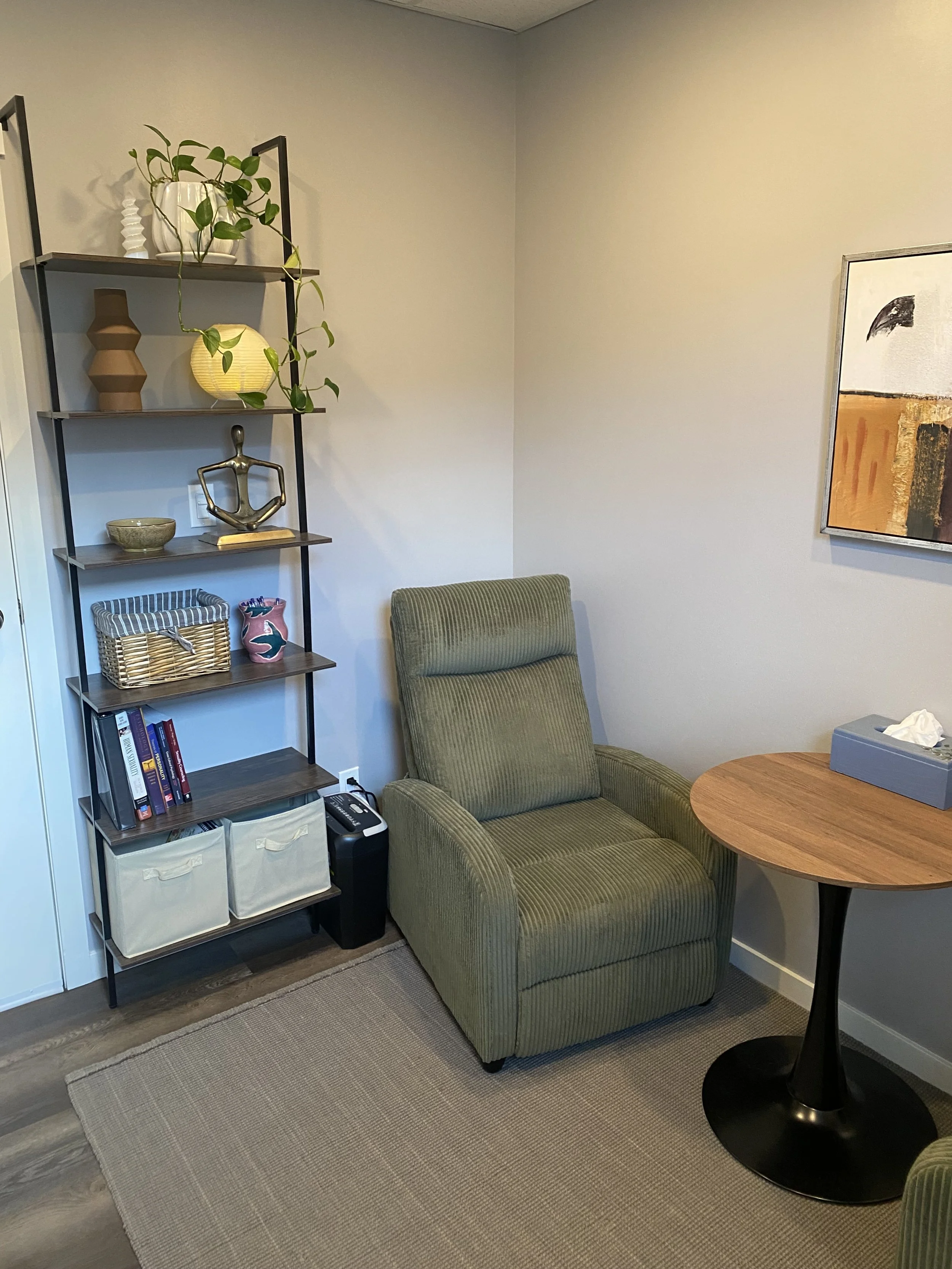 A cozy therapy office with a green upholstered armchair, a round wooden table, a black metal bookshelf with decorative items, a woven basket, and a framed abstract artwork on a gray wall. A beige rug covers part of the dark wood flooring.