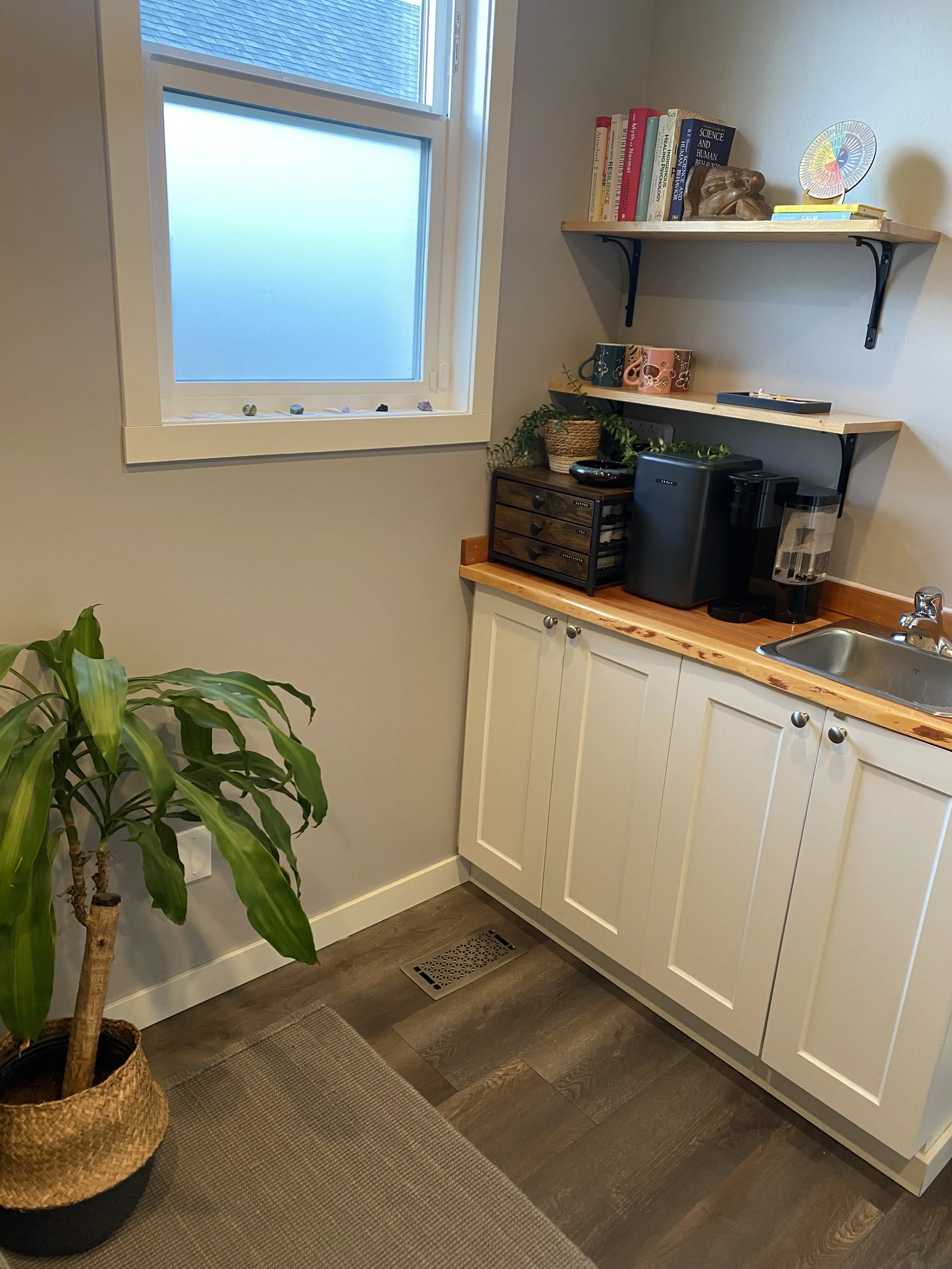 A therapy office corner with a coffee bar, window, white cabinets, wooden countertops, a plant in a woven basket, and floating shelves with books and decorative items.