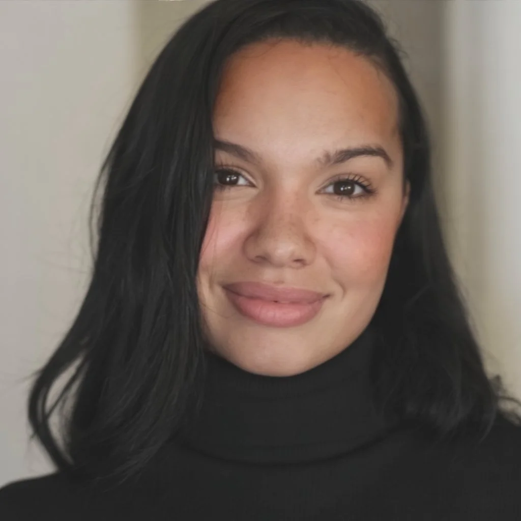 Close-up of a woman with long black hair, smiling, wearing a black turtleneck, standing indoors with a neutral background.