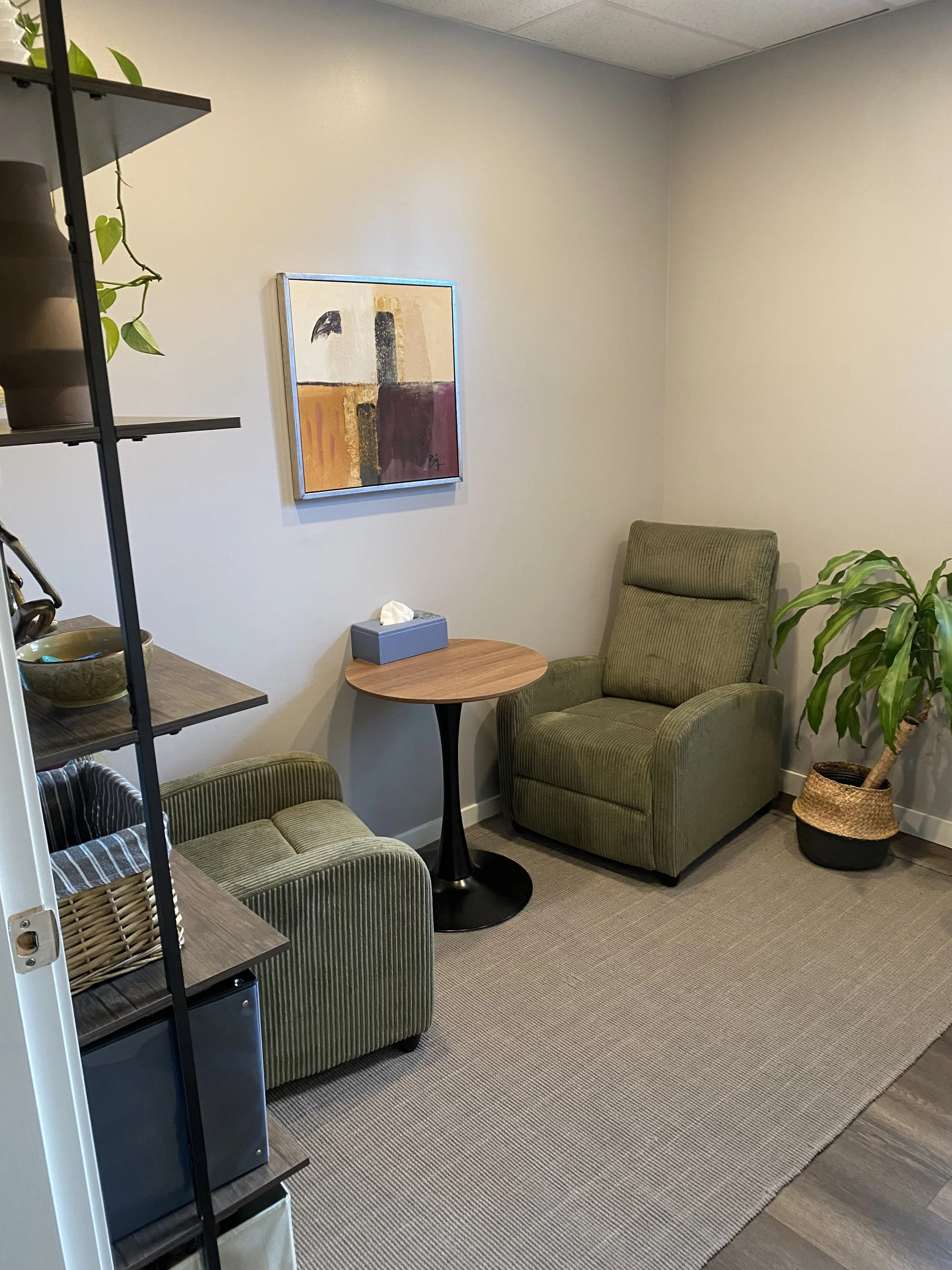 A cozy office corner with two green armchairs, a small round wooden table, a tall potted plant, a bookshelf with decorative items, and a framed abstract painting on the wall.