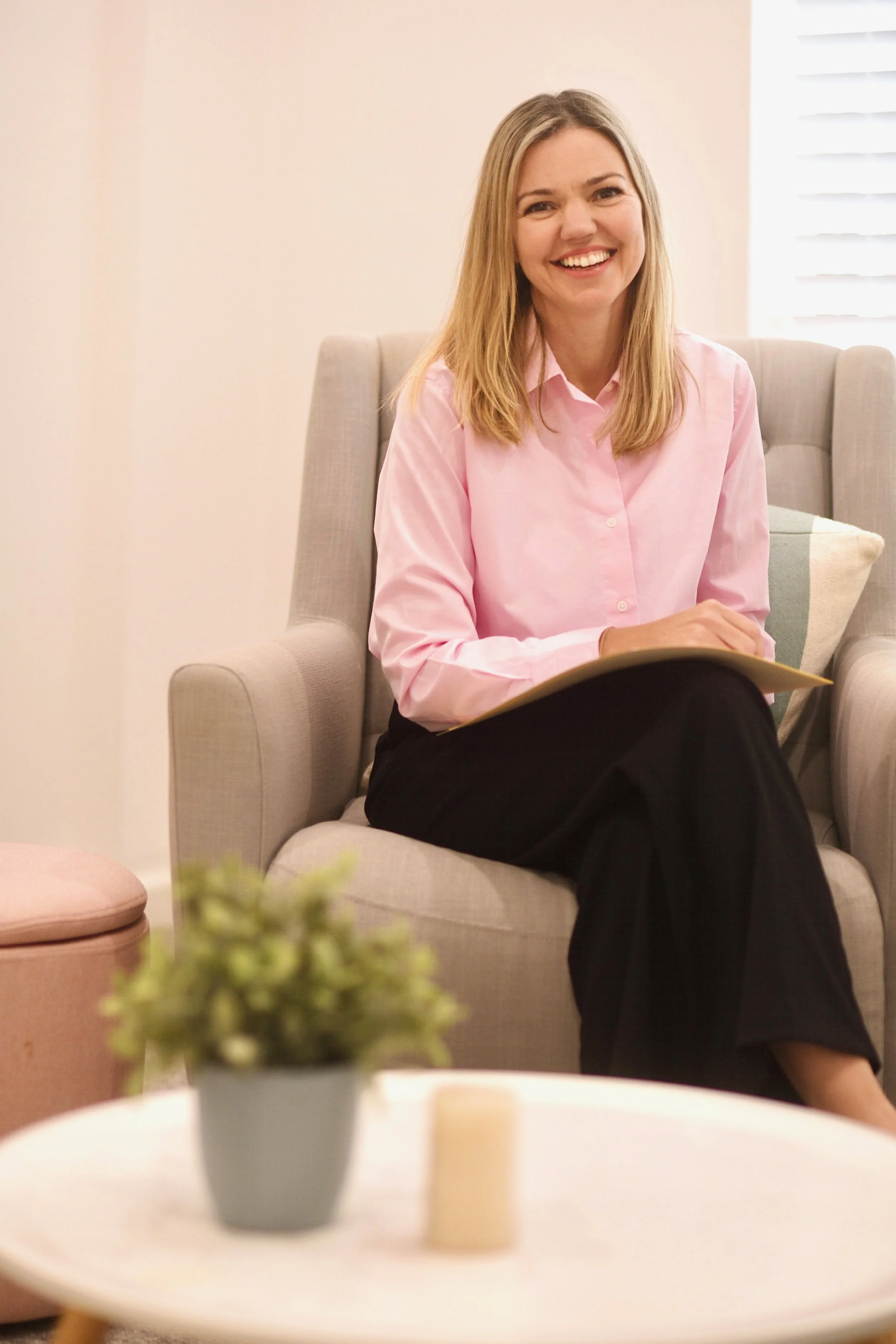 A smiling woman in a pink shirt and black pants sitting on a light-colored armchair in a bright room with white blinds. In the foreground, a round table with a small potted plant.
