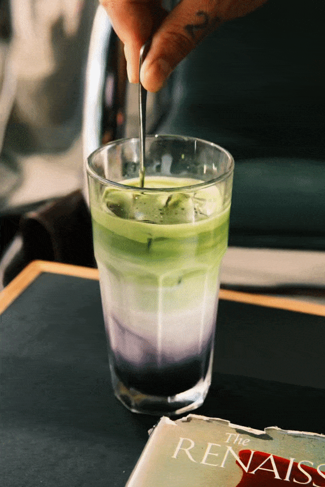 A layered colorful beverage in a tall glass with a metal stirrer being inserted, placed on a dark table with a book titled 'The Renaissance' nearby.