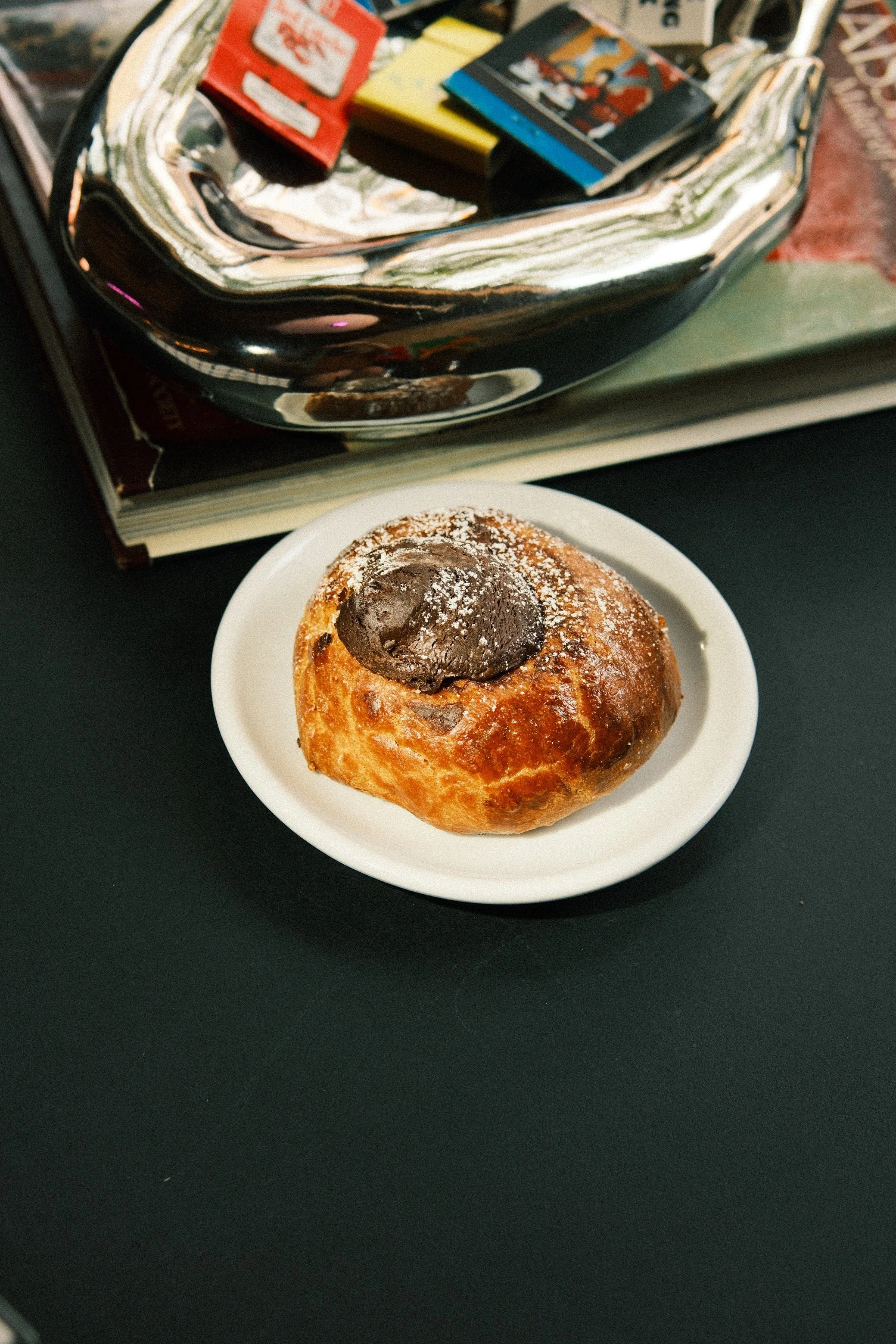 A small plate with a chocolate chip muffin topped with a dollop of chocolate and powdered sugar, placed on a black surface. In the background, a book and snacks are visible.